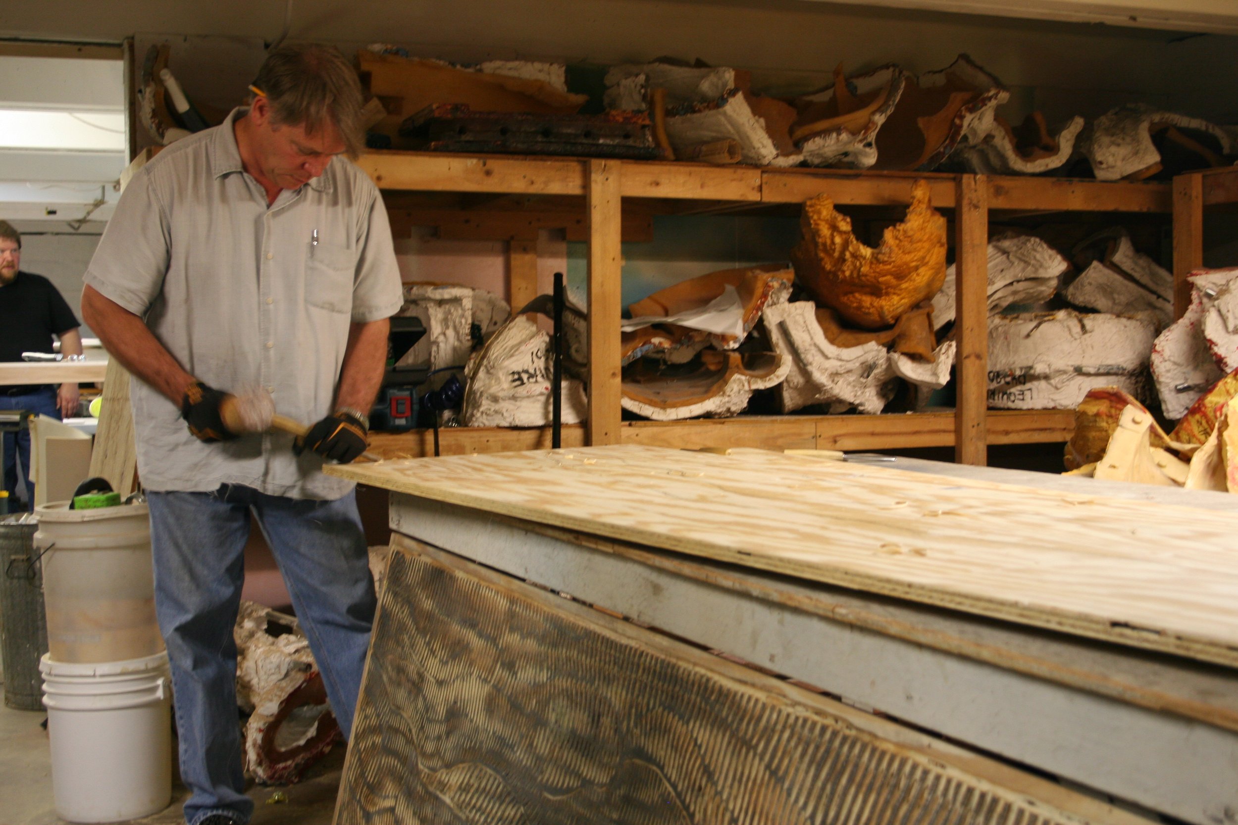 A man working with wood in a workshop with shelves of various wood pieces in the background.