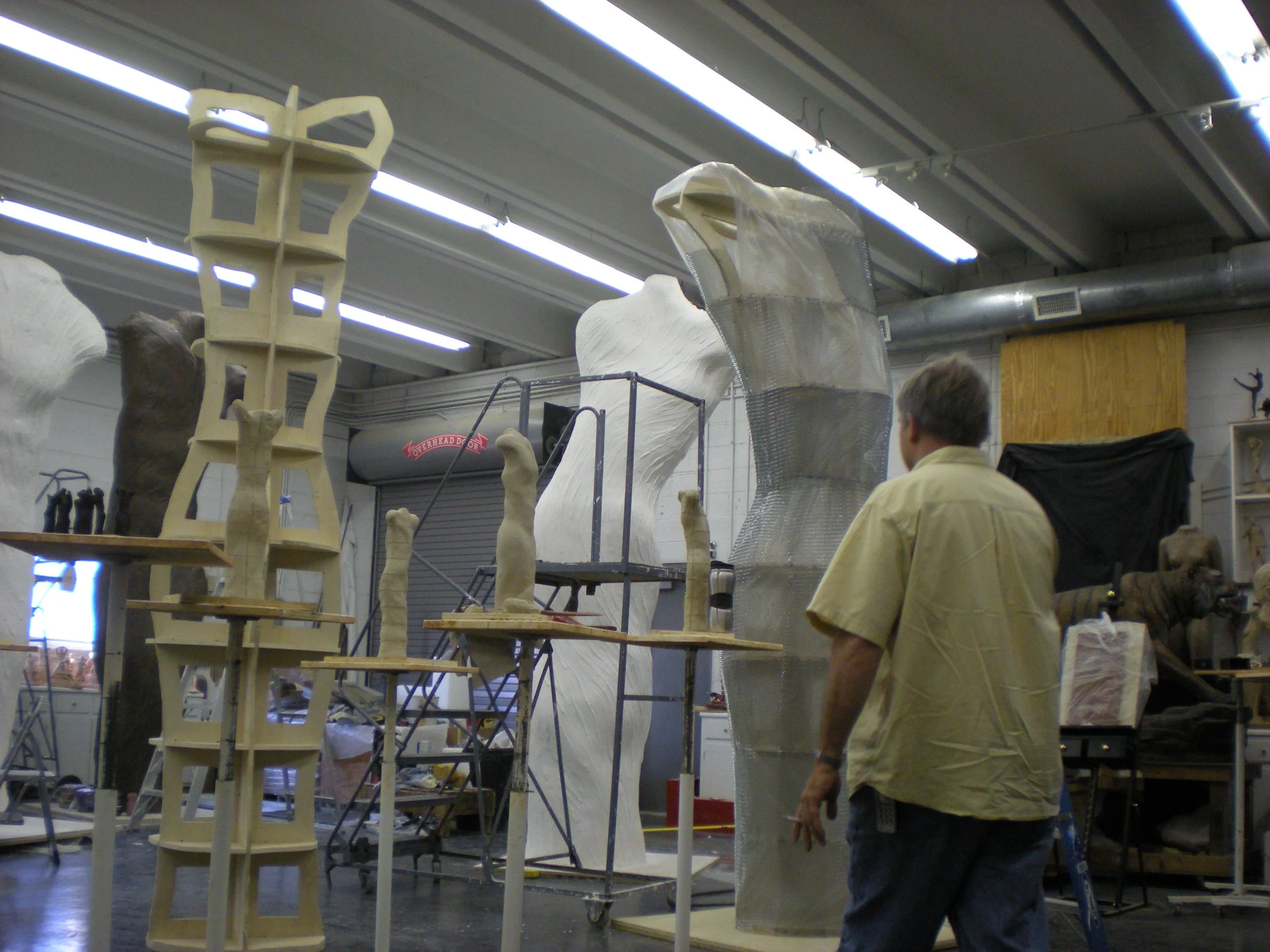 Workshop with sculptures and models, including a wooden armature, a partially covered large sculpture, and various tools and materials on tables and shelves.