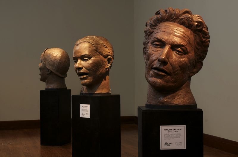 Three bronze bust sculptures of women, displayed on black pedestals inside a gallery. The right bust is labeled 'Woody Guthrie,' depicting an older man with textured hair.  Cultural Heroes