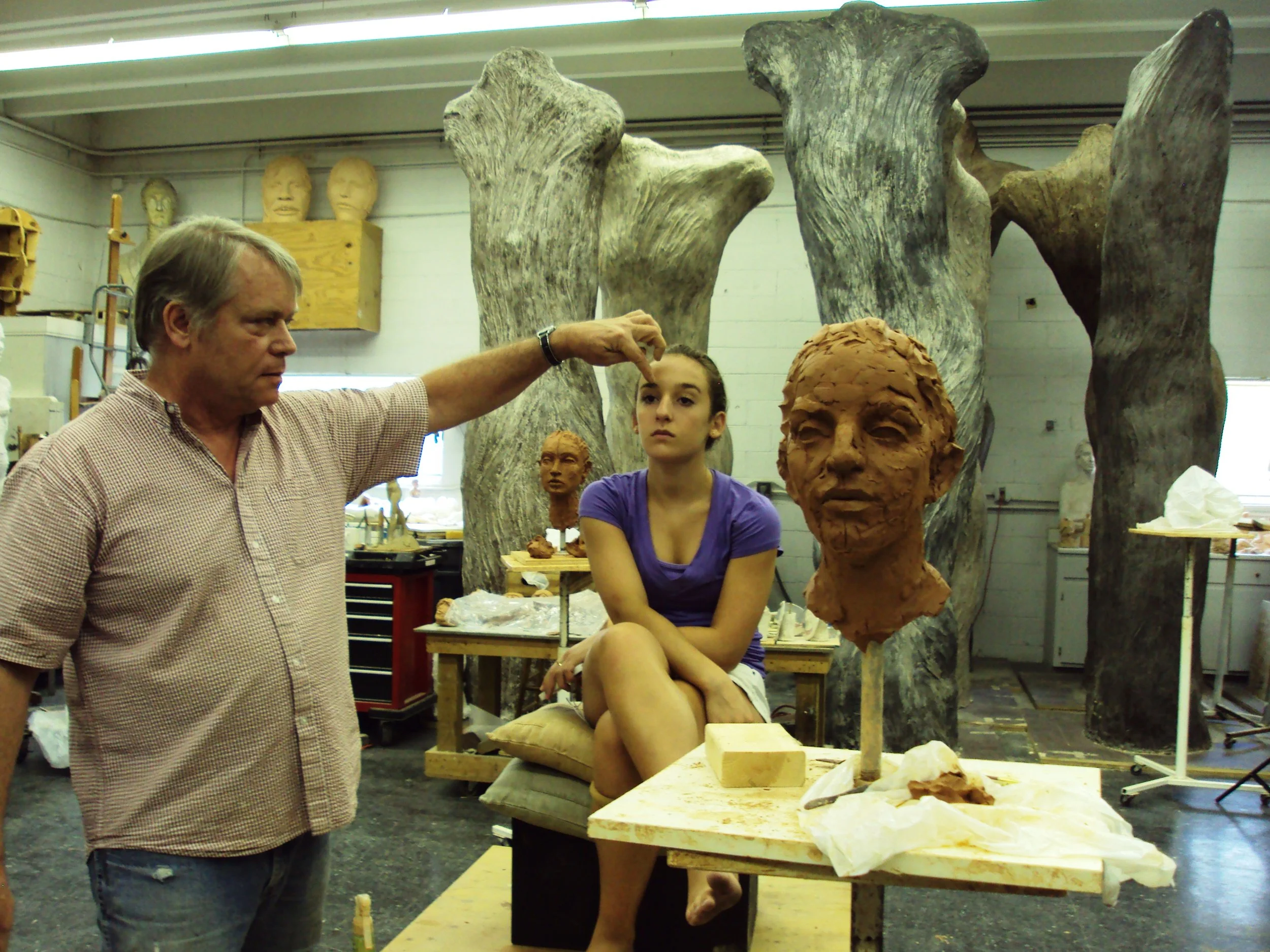 An older man and a young woman working on sculptures in a workshop, surrounded by unfinished busts and large wooden molds.