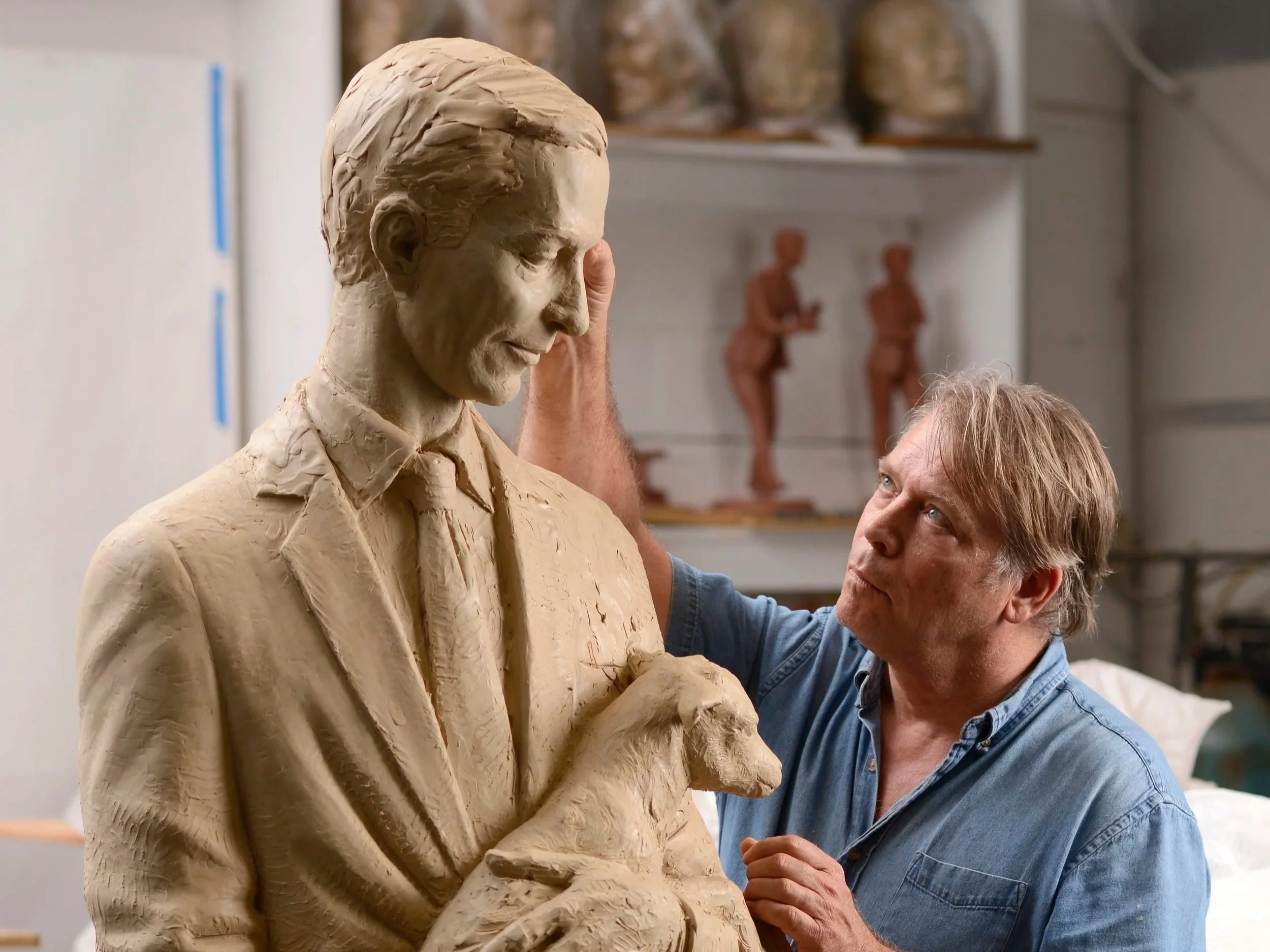 An artist shaping a clay sculpture of a woman with short hair, wearing a blazer, in a studio. The artist is a man with glasses and a blue denim shirt, focused on his work.