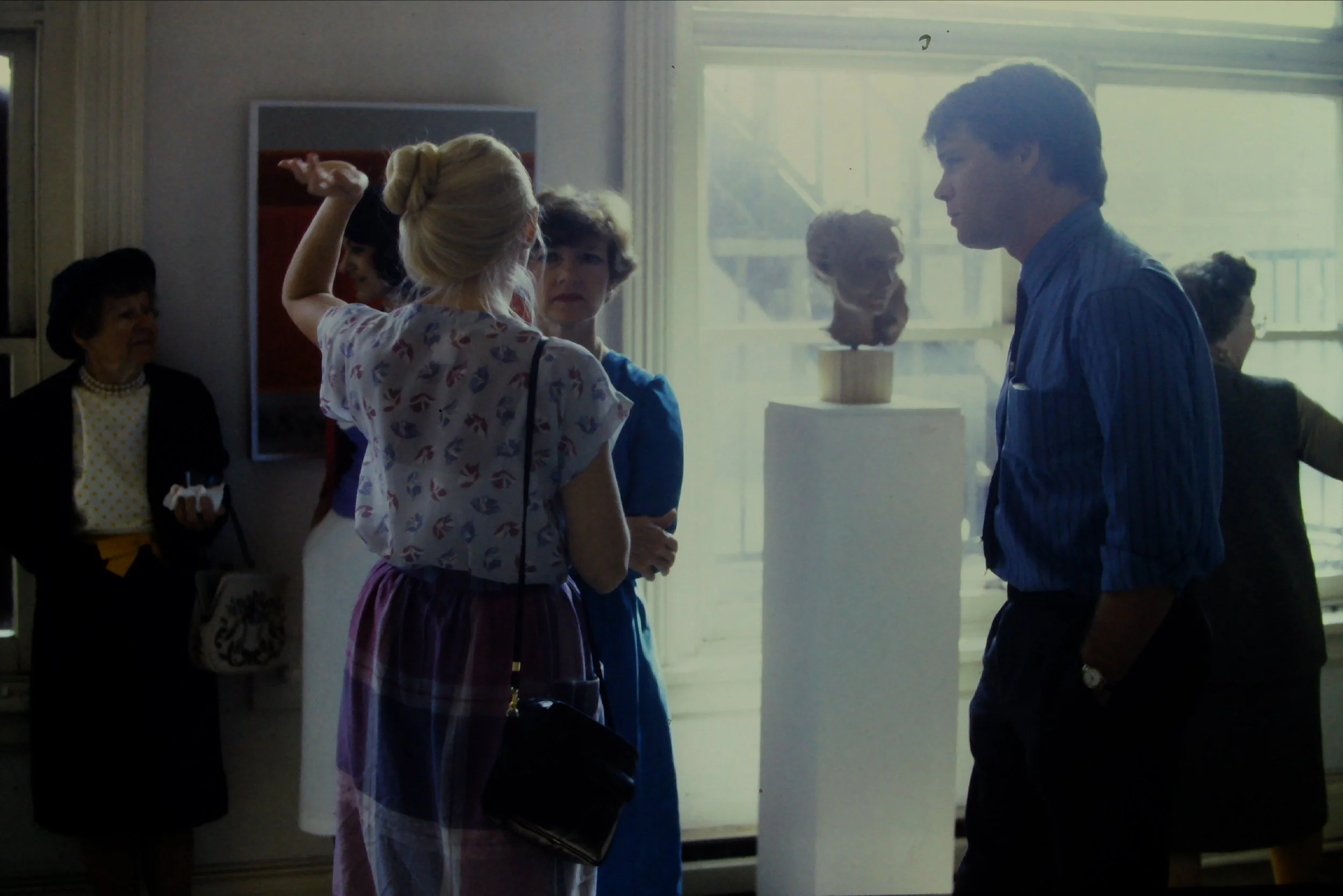 A group of people inside a building, engaging in conversation near a large window with a sculpture of a head on a stand in the background.