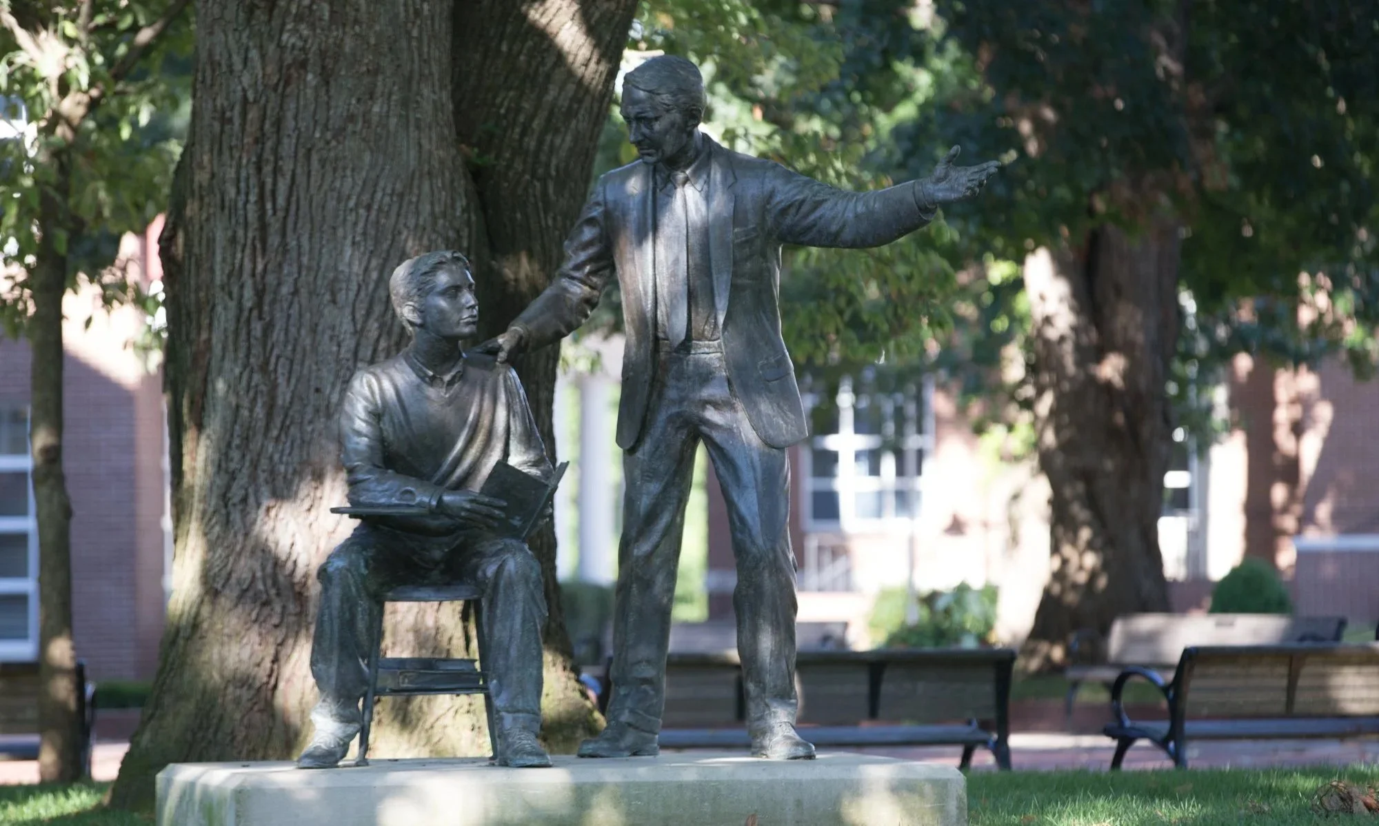 Bronze statue of a man in a suit gesturing with his right arm, and a woman sitting on a chair reading a book, set outdoors with trees and benches in the background.