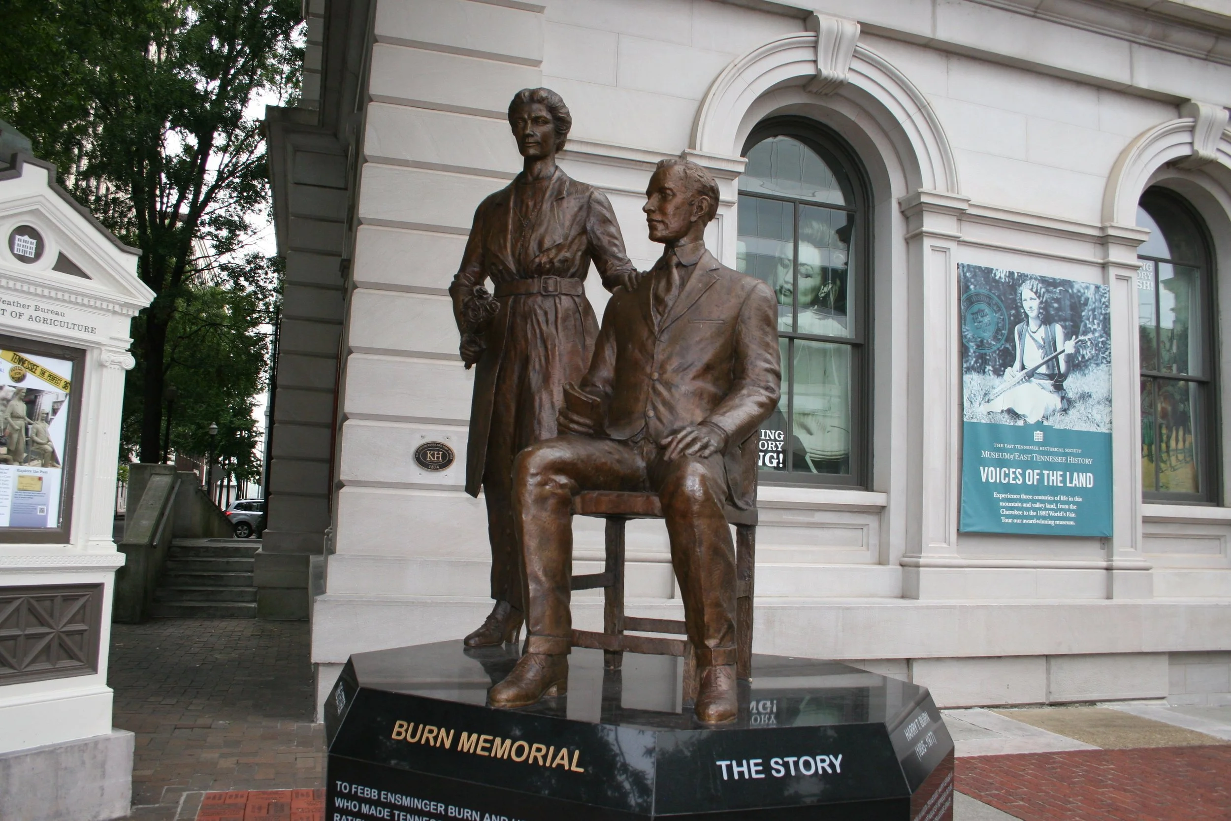 Bronze sculpture of a seated man holding a book, with a standing woman beside him, in front of a historical building with banners about the Tennessee history museum.