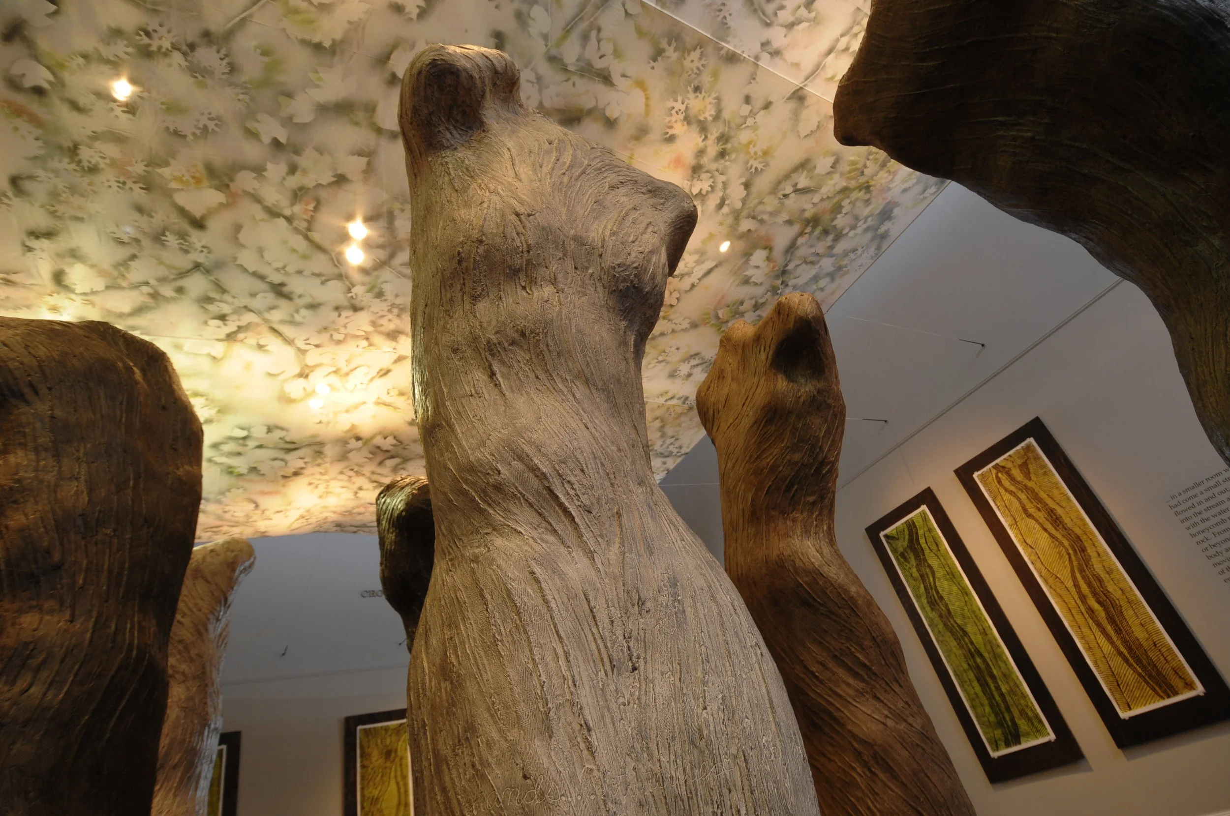 Multiple large wooden sculptures resembling tree trunks are displayed indoors on a boardwalk. In the background, there are framed art pieces on a wall and a ceiling decorated with floral patterns and lighting. Dream Forest Installation. 