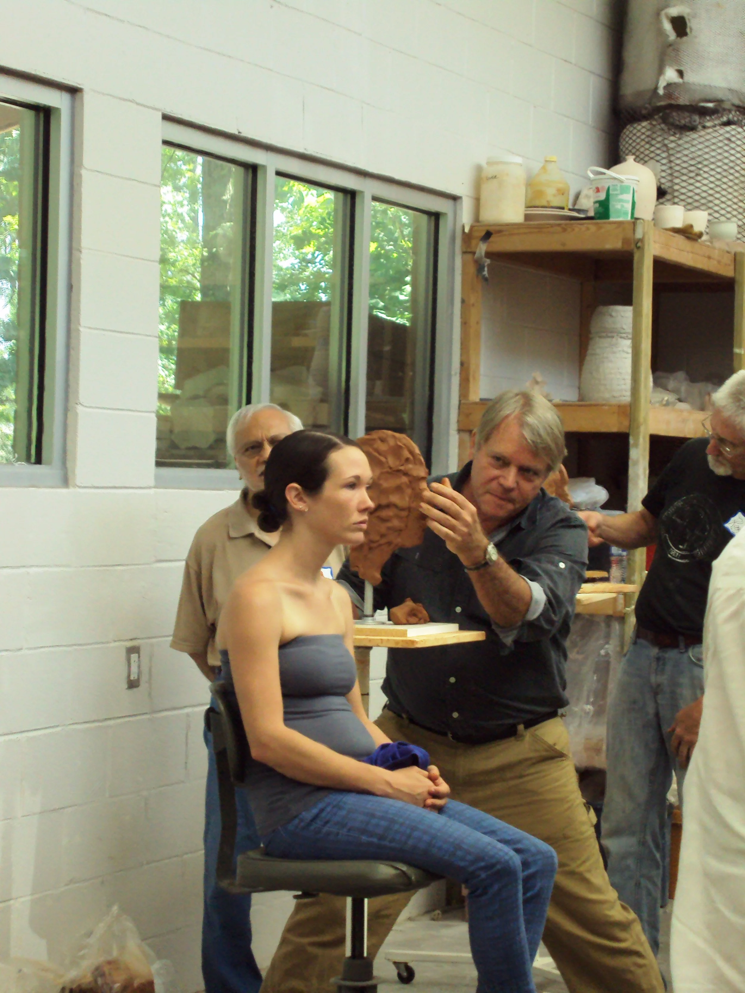 People working on a clay sculpture in an art studio. One woman seated with a sculpture on her lap, surrounded by others engaged in the process.