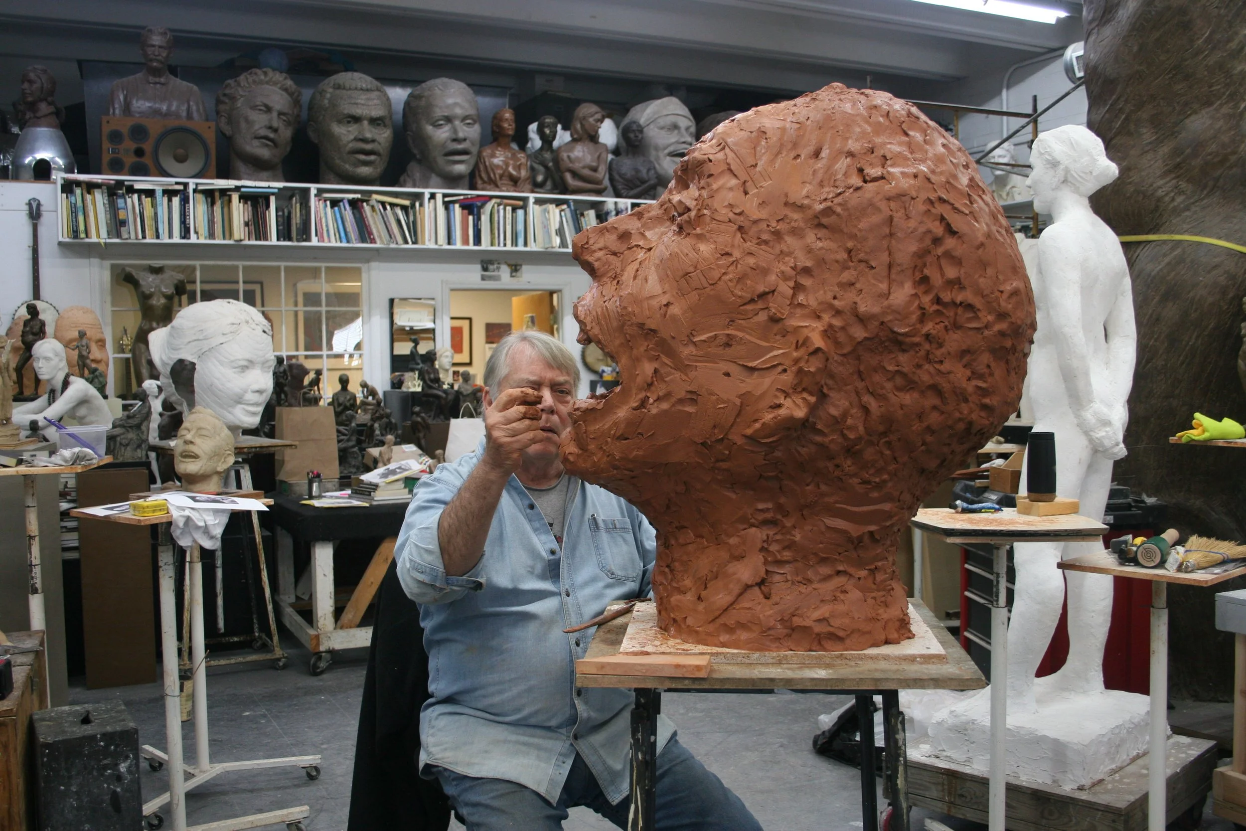 An artist working on a large clay sculpture of a person's head Pete Seeger in an art studio with busts and sculptures on shelves in the background.
