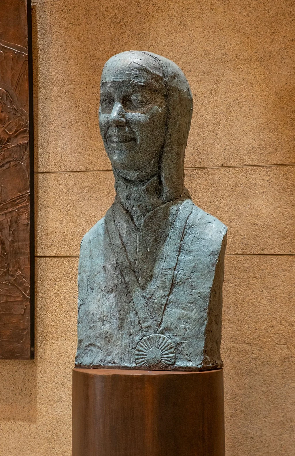 A plaster bust sculpture of a woman wearing a medal necklace with a radiating sun design, placed on a metal pedestal against a beige wall.