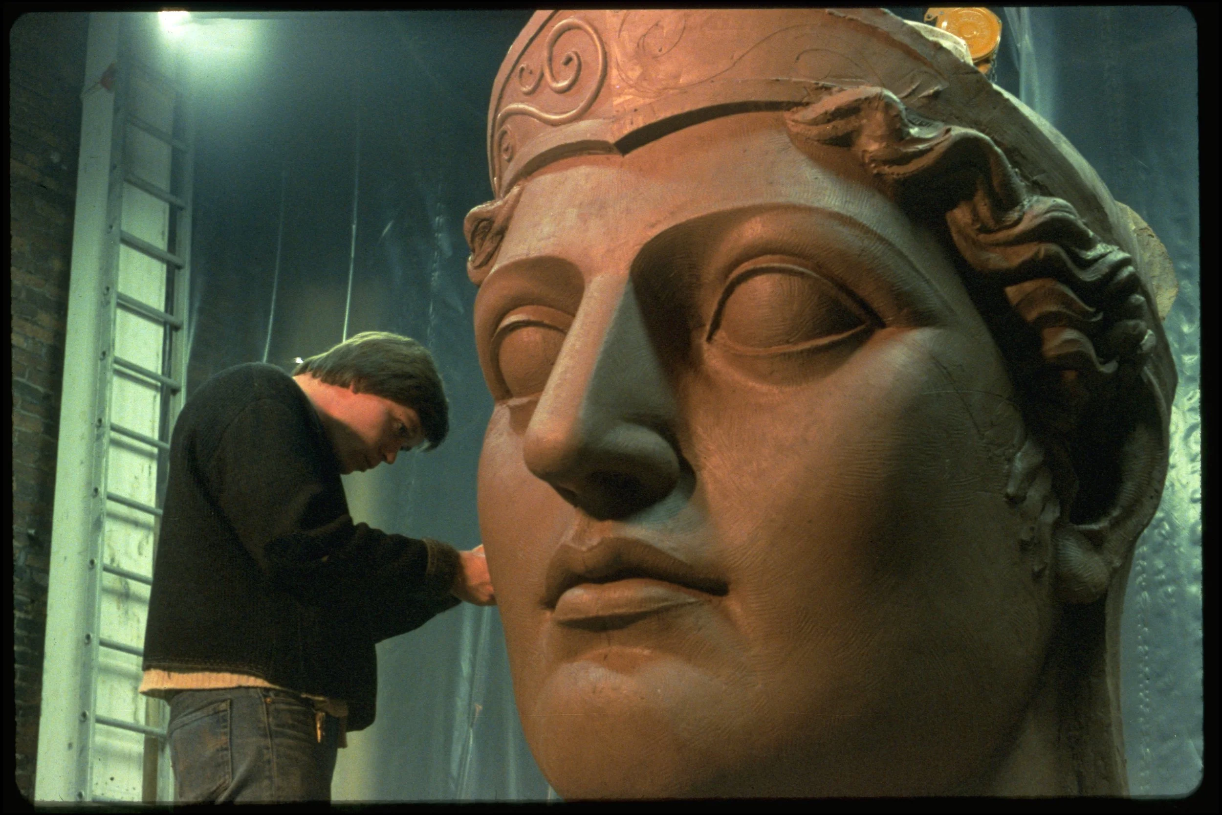 Person working on a large clay sculpture of a Athenas face, wearing a black sweater and gray pants, with a ladder and workshop background. Athena Parthenos in Parthenon Museum Nashville. 