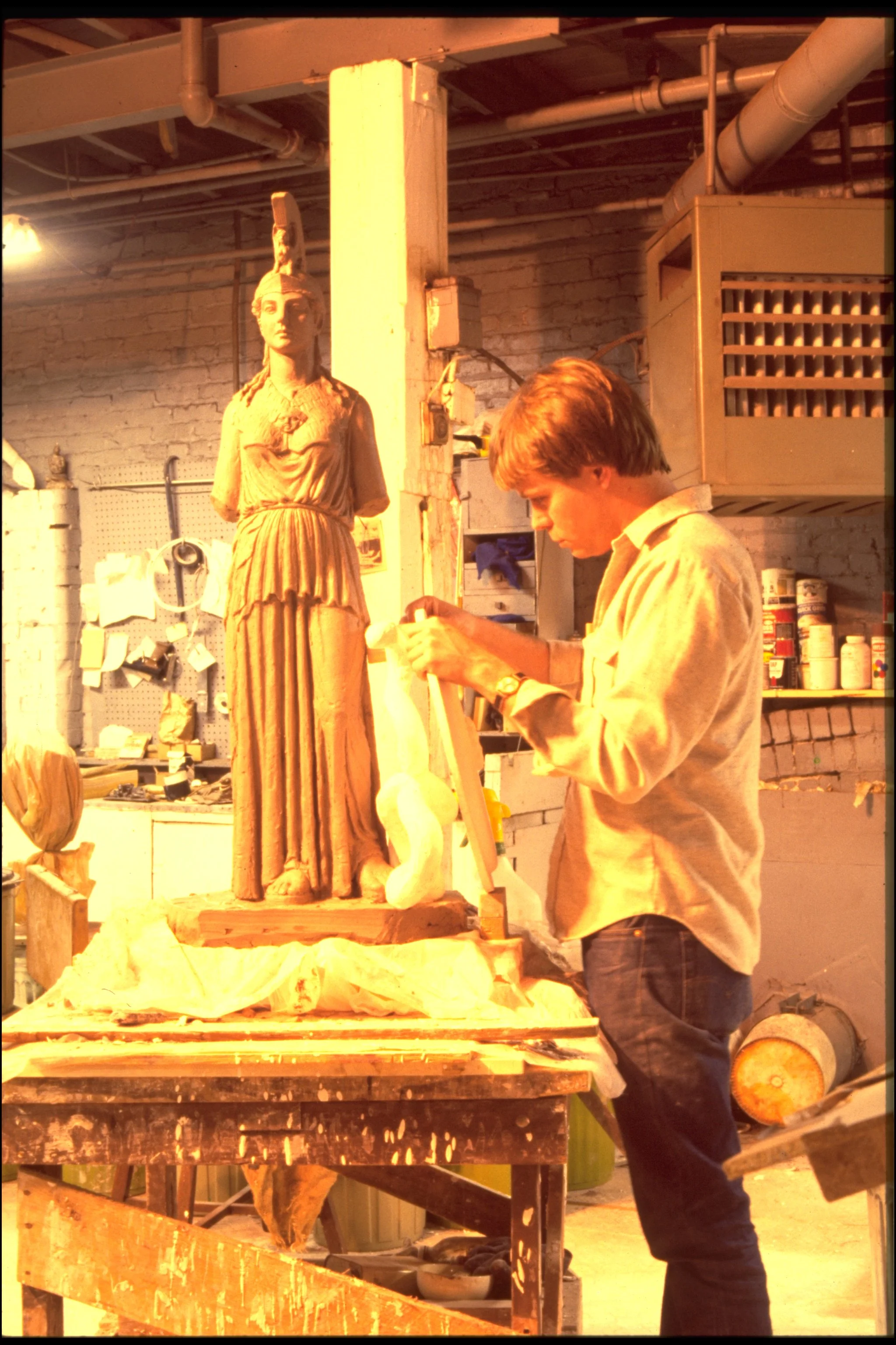 A young man is sculpting a clay statue of a woman with a detailed dress and a headdress in an art studio with brick walls, tools, and shelves in the background. Athena Parthenos in Parthenon Museum Nashville. 