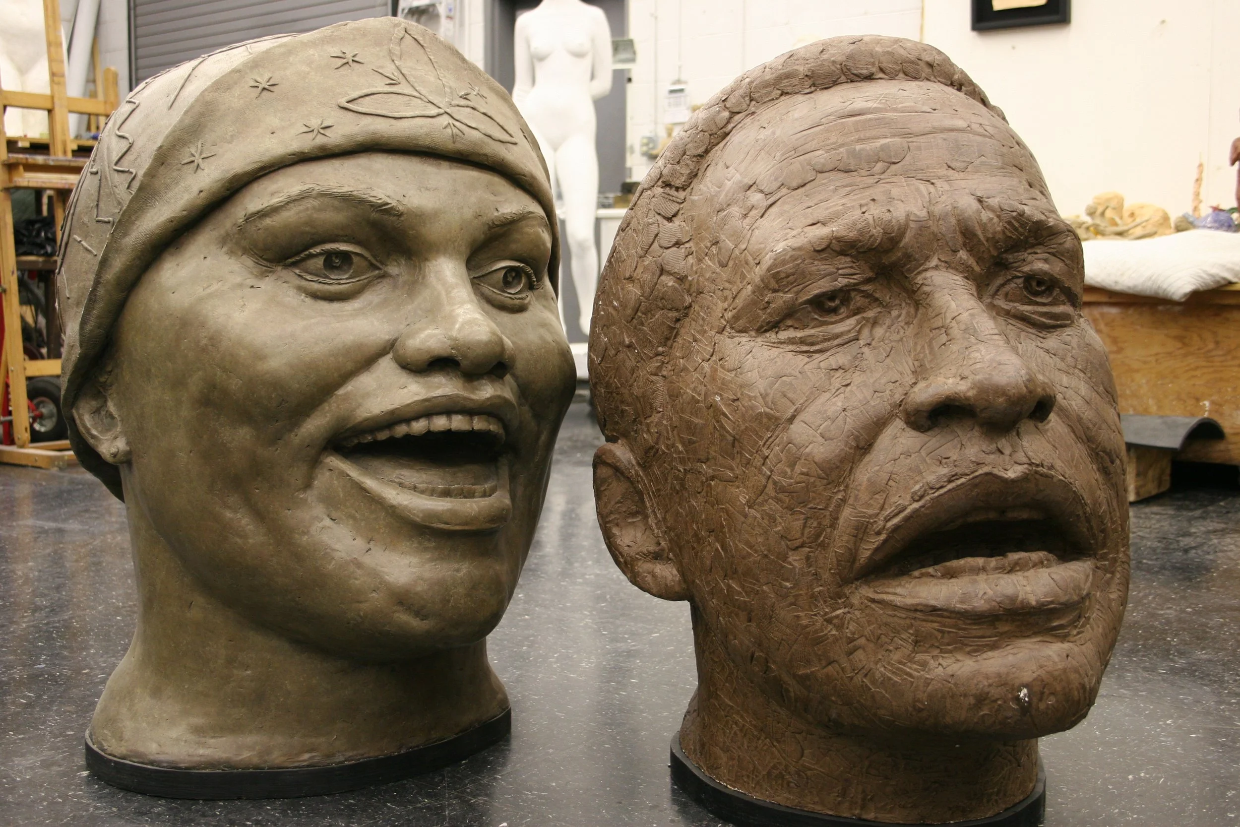 Two large sculpture busts on display in a studio. One is a realistic face of a woman wearing a headscarf with detailed facial features. The other is a stylized face of a person with exaggerated features, carved from clay.  Cultural Heroes