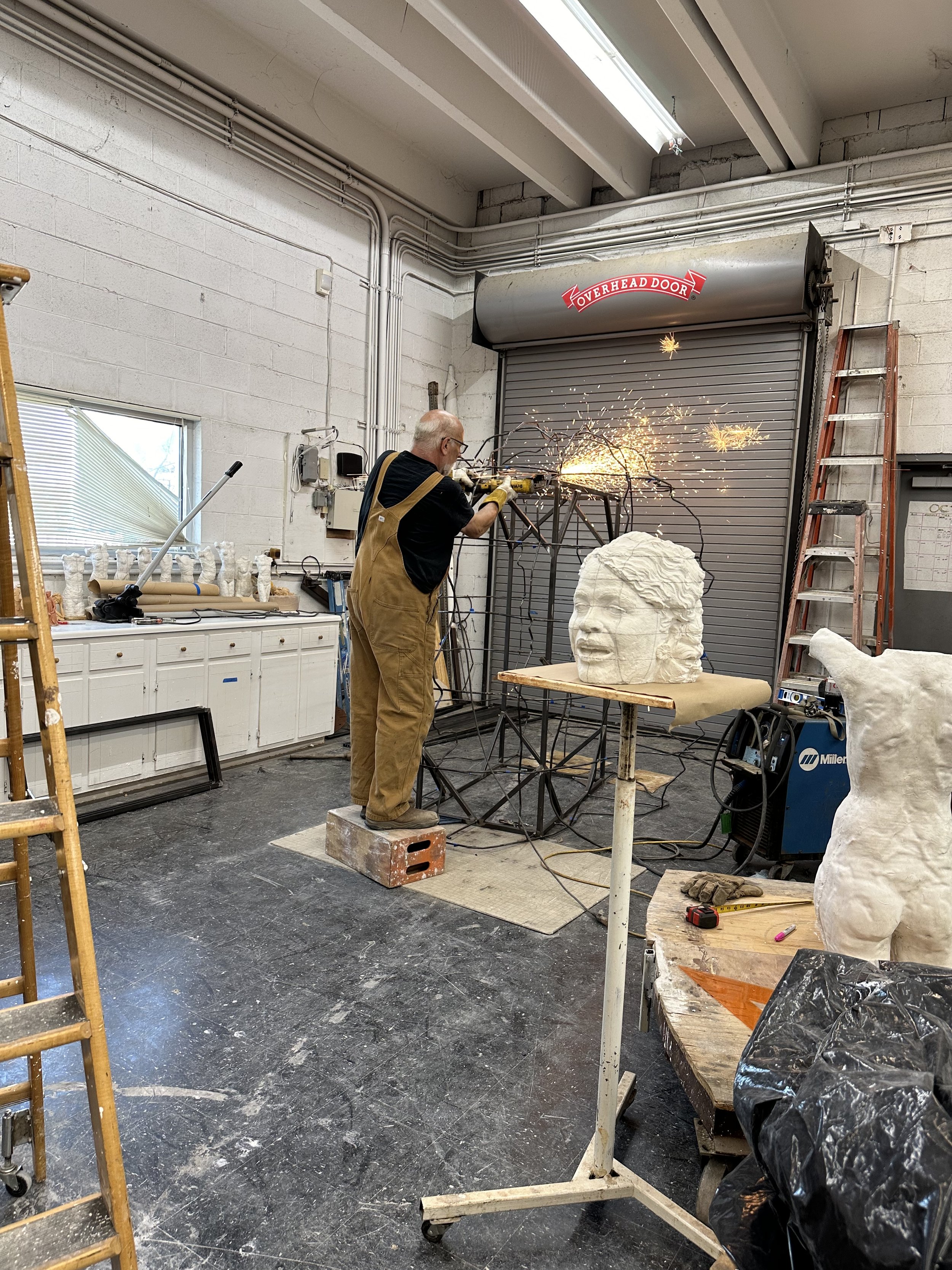 A man working on a metal sculpture in a workshop, welding sparks flying, with a large head sculpture on the table, surrounded by various tools and equipment, including a ladder, workbench, and a rolling door labeled 'Overhead Door'.