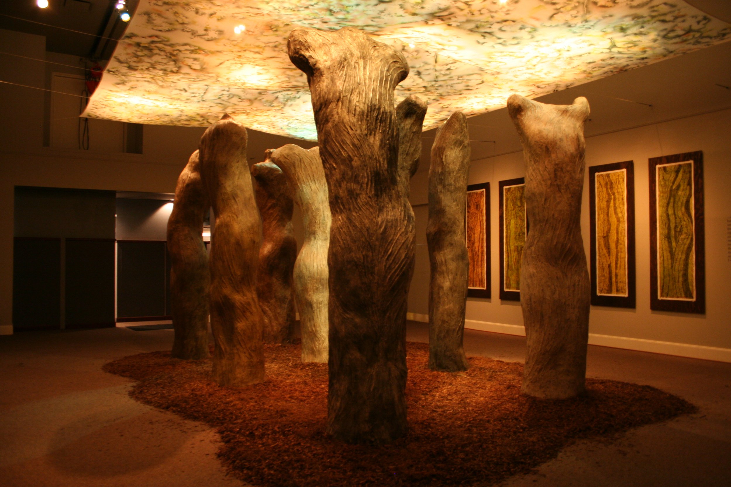 An art installation or sculpture exhibition featuring large, abstract, organic wooden forms resembling tree trunks, with a colorful, illuminated ceiling overhead. Framed artwork is visible on the wall in the background. Dream Forest Installation.
