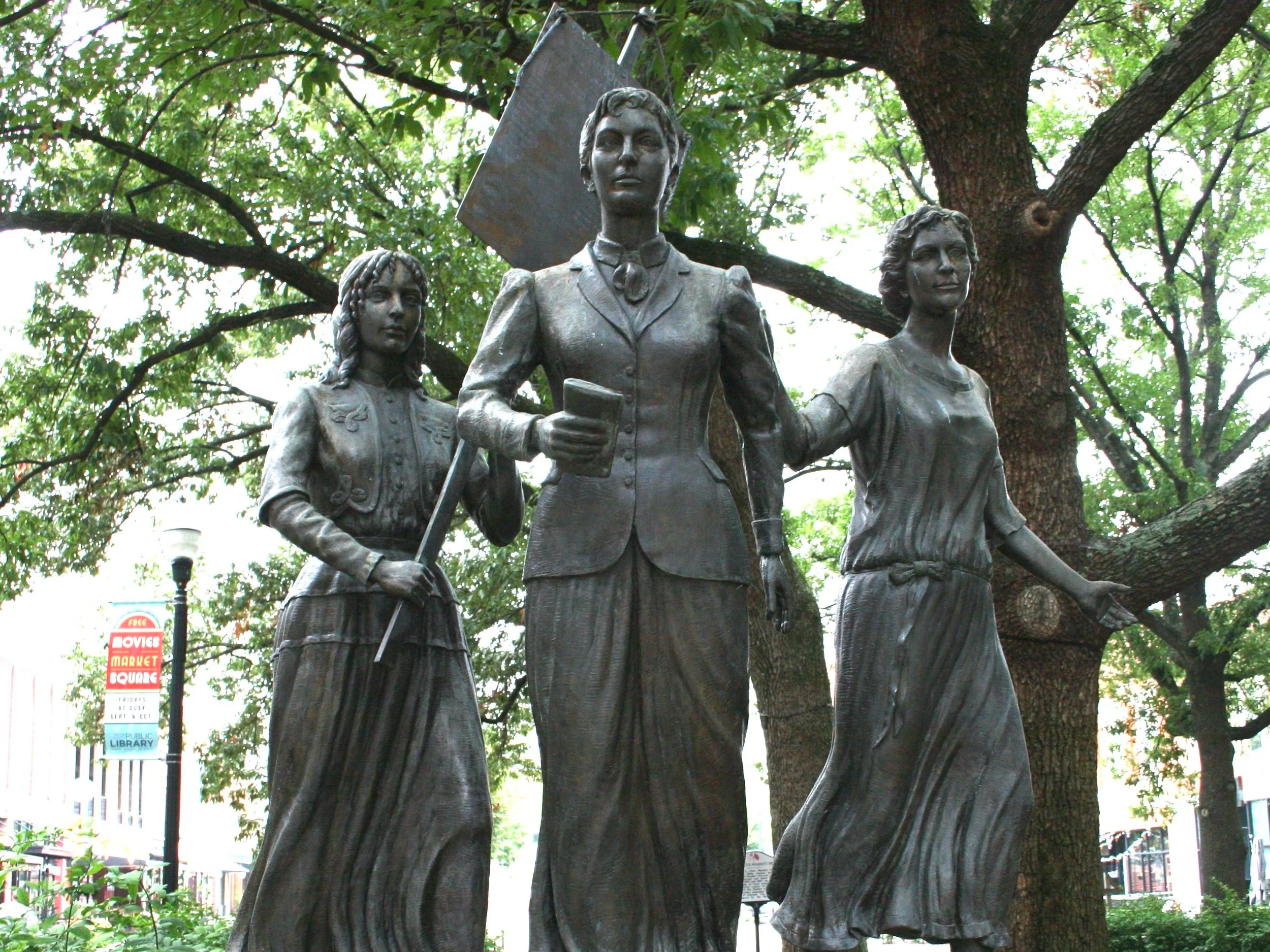 Statue of three women holding over each other, standing outdoors under a large tree.