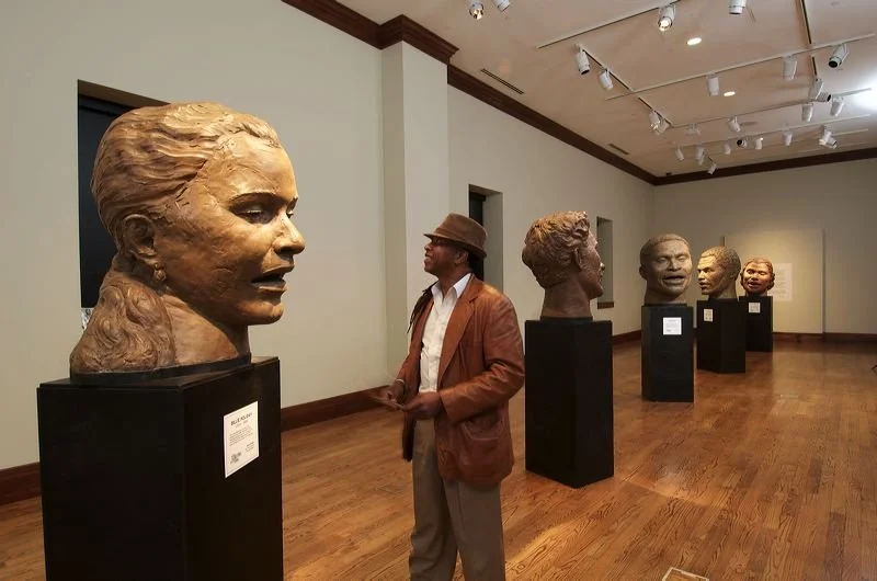 A man in a brown jacket, beige trousers, and a brown hat stands in an art gallery, observing a large bronze bust of a woman. The gallery features multiple bust sculptures on black pedestals, with a wooden floor and white walls.  Cultural Heroes