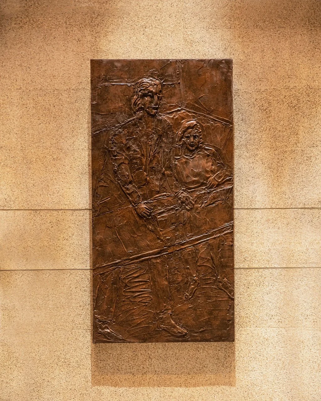 Bronze relief sculpture of two people, a woman and a child, woman the teacher holding a book, mounted on a beige wall.