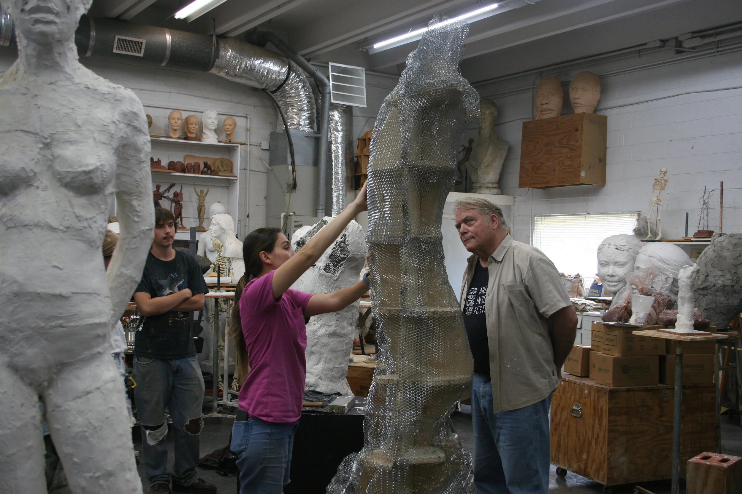 Alan Lequire and Amber Lelli in an art studio working on a sculpture, with model heads and other sculptures in the background.