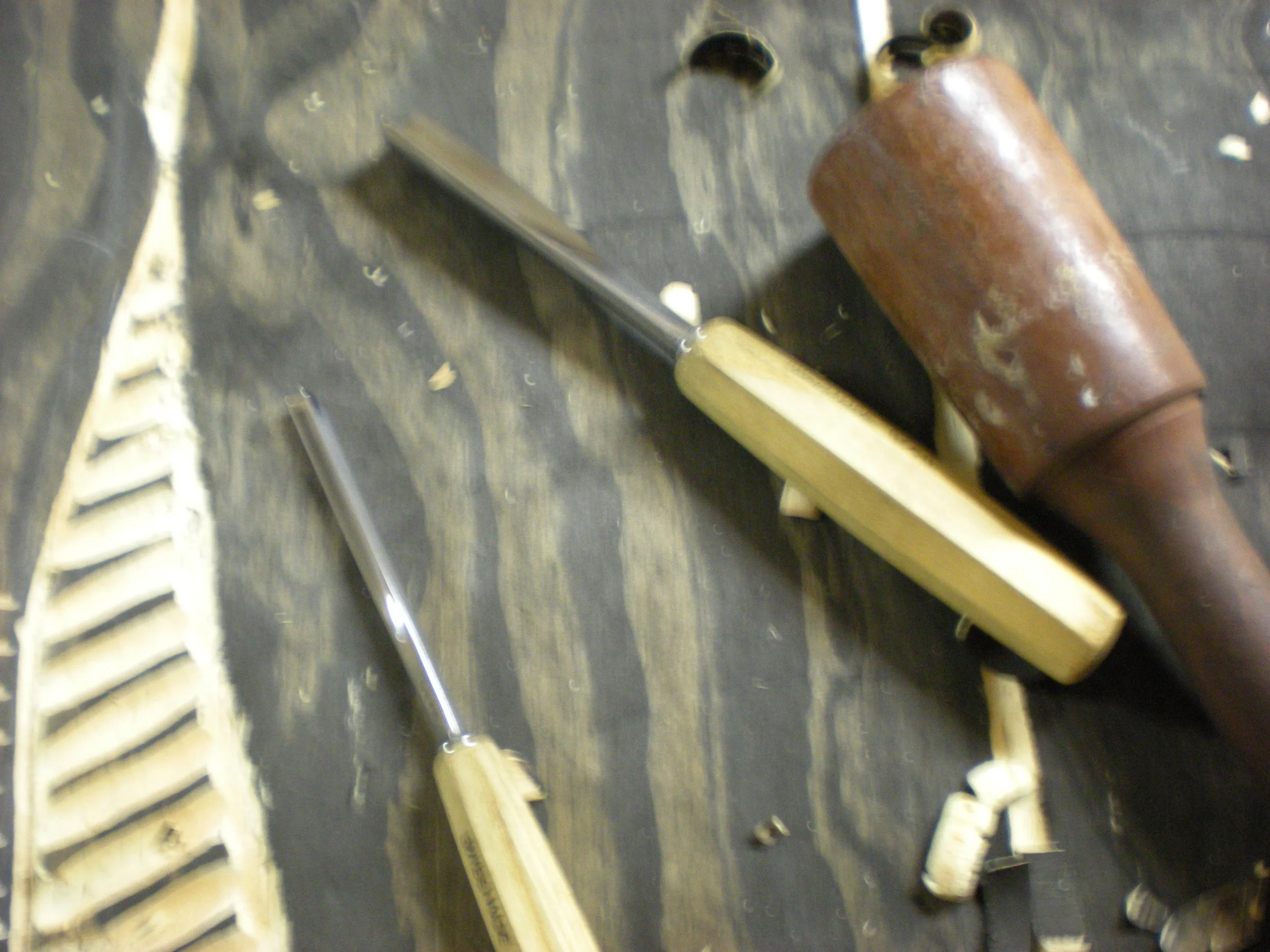 The image shows several tools and parts scattered on a dark work surface, including a mallet with a brown wooden handle, a metal chisel, and elongated wooden or metal pieces.