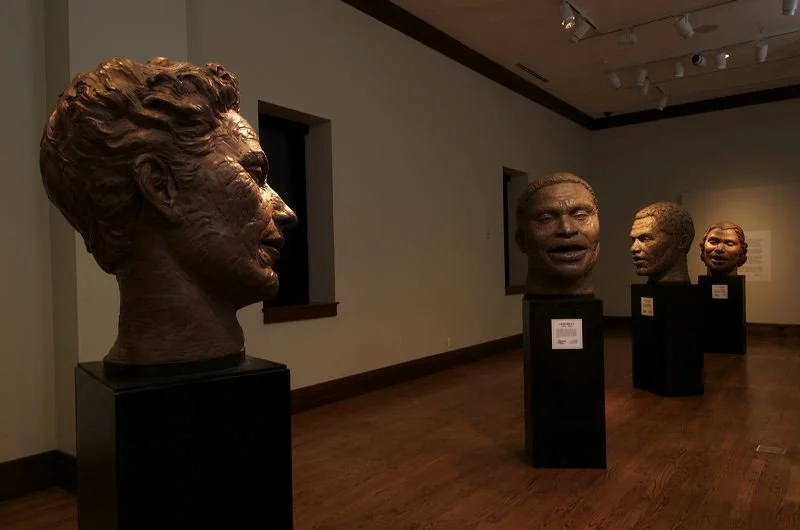 Four sculpted busts of diverse people's heads displayed on black pedestals in an art gallery.  Cultural Heroes