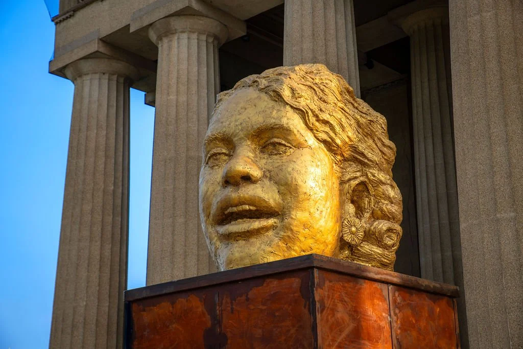 A large golden bust of a woman on a wooden pedestal, in front of a building with tall columns.