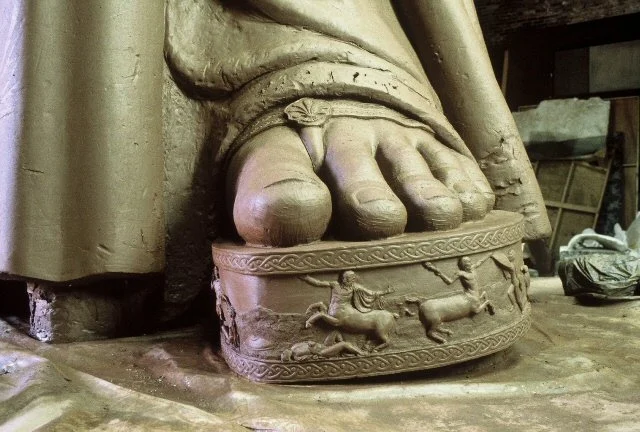 Close-up of a clay sculpture of a large statue or sculpture, featuring detailed reliefs of animals and patterns, with the bottom of a person's foot resting on it. Athena Parthenos in Parthenon Museum Nashville. 