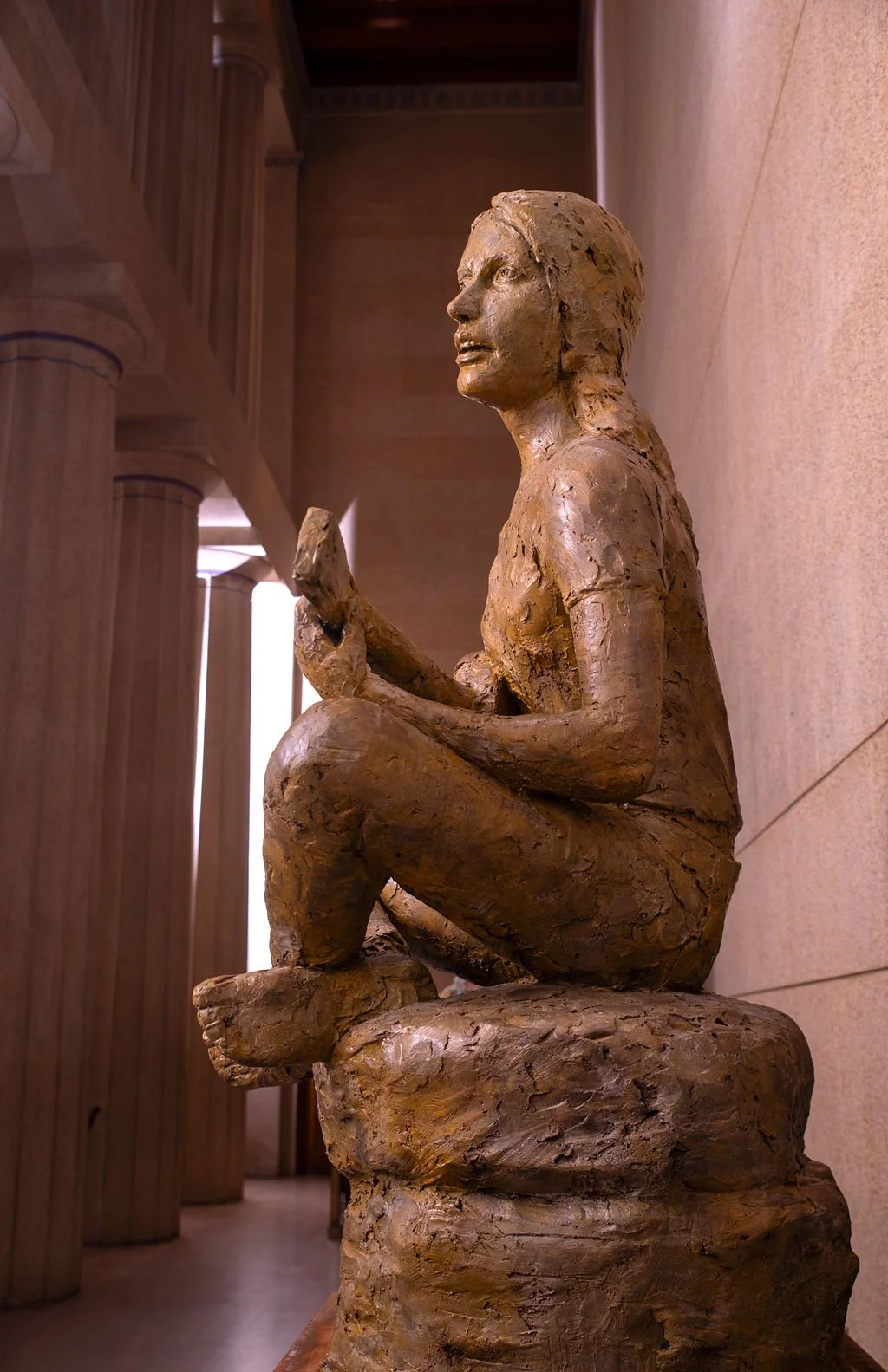 Bronze sculpture of a singer Joan Baez woman kneeling with one knee on a rock, holding an object in her hands, inside a building with columns.