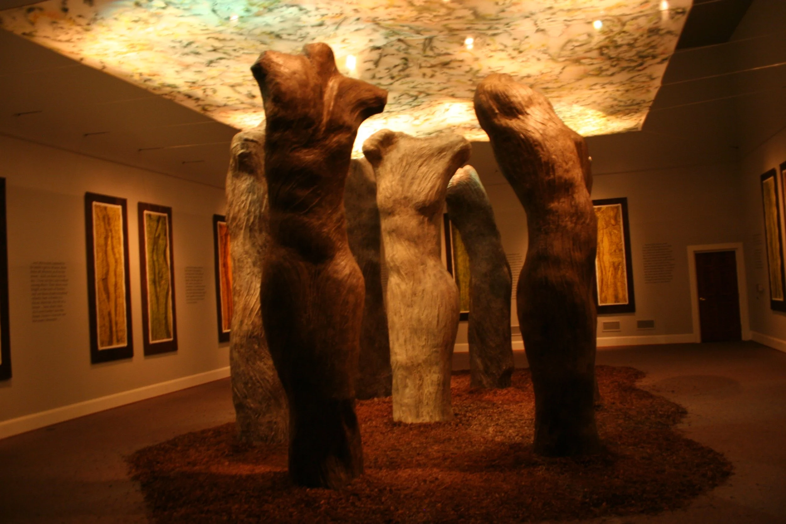 Inside a museum or gallery, large wooden sculptures of abstract human figures standing in a row. The ceiling has colorful, patterned lighting, and framed artwork hangs on the wall. Dream Forest Wood Block Prints.