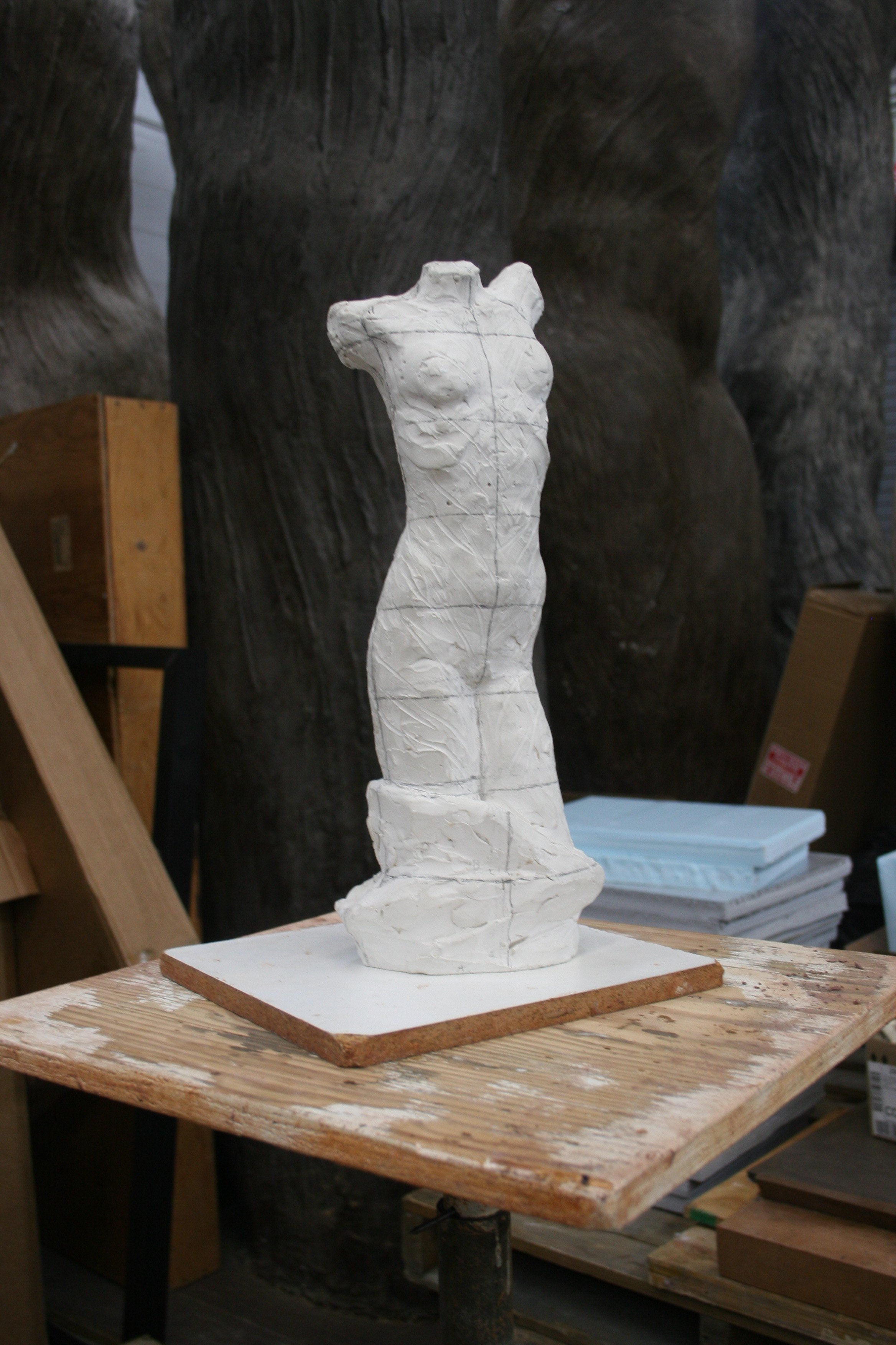 A white sculpture of a female torso on a wooden base, placed on a worktable in an art studio.