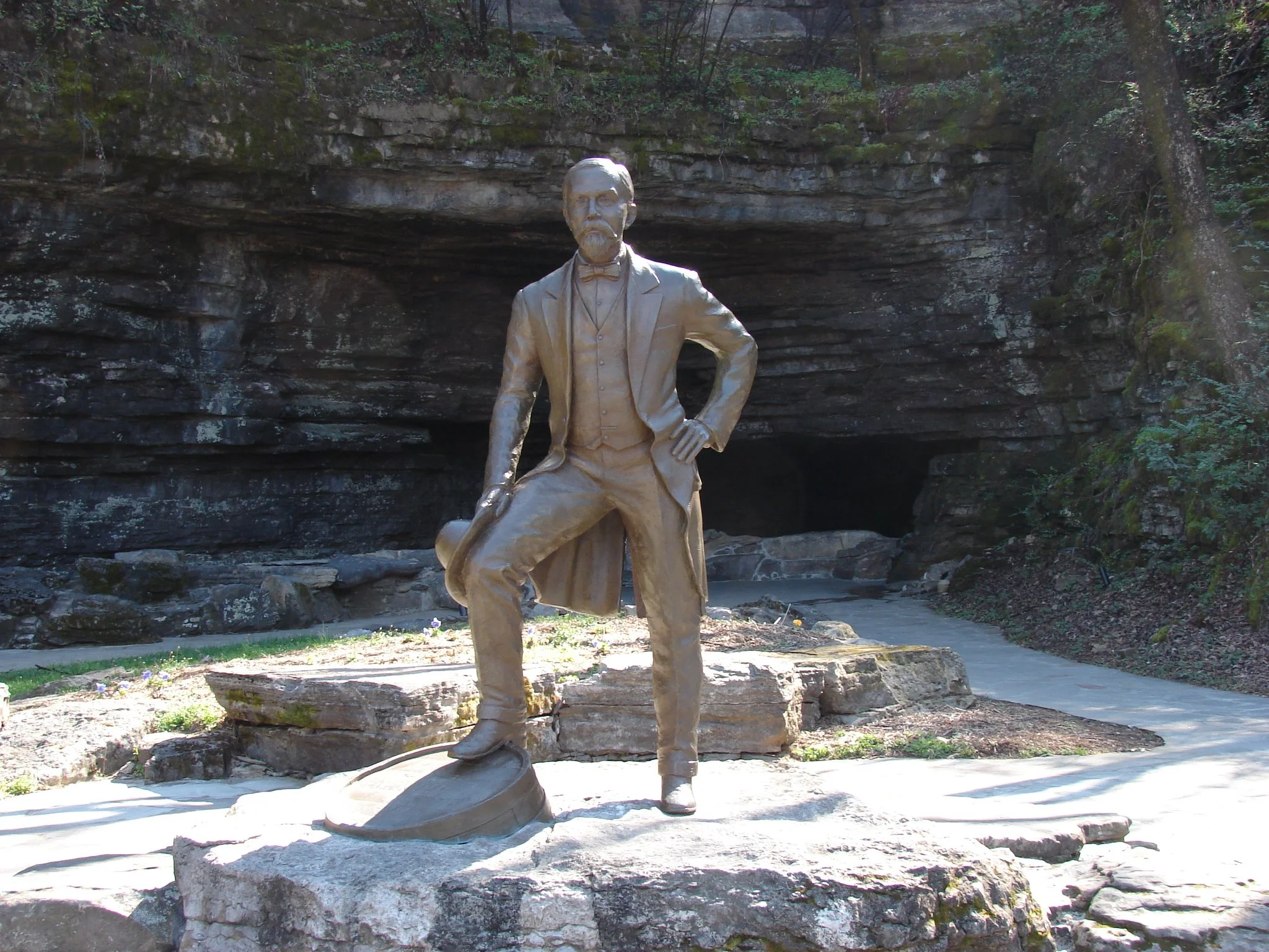 Bronze statue of a man in a suit, standing with one hand on his hip, in front of a rocky cliff and trees.