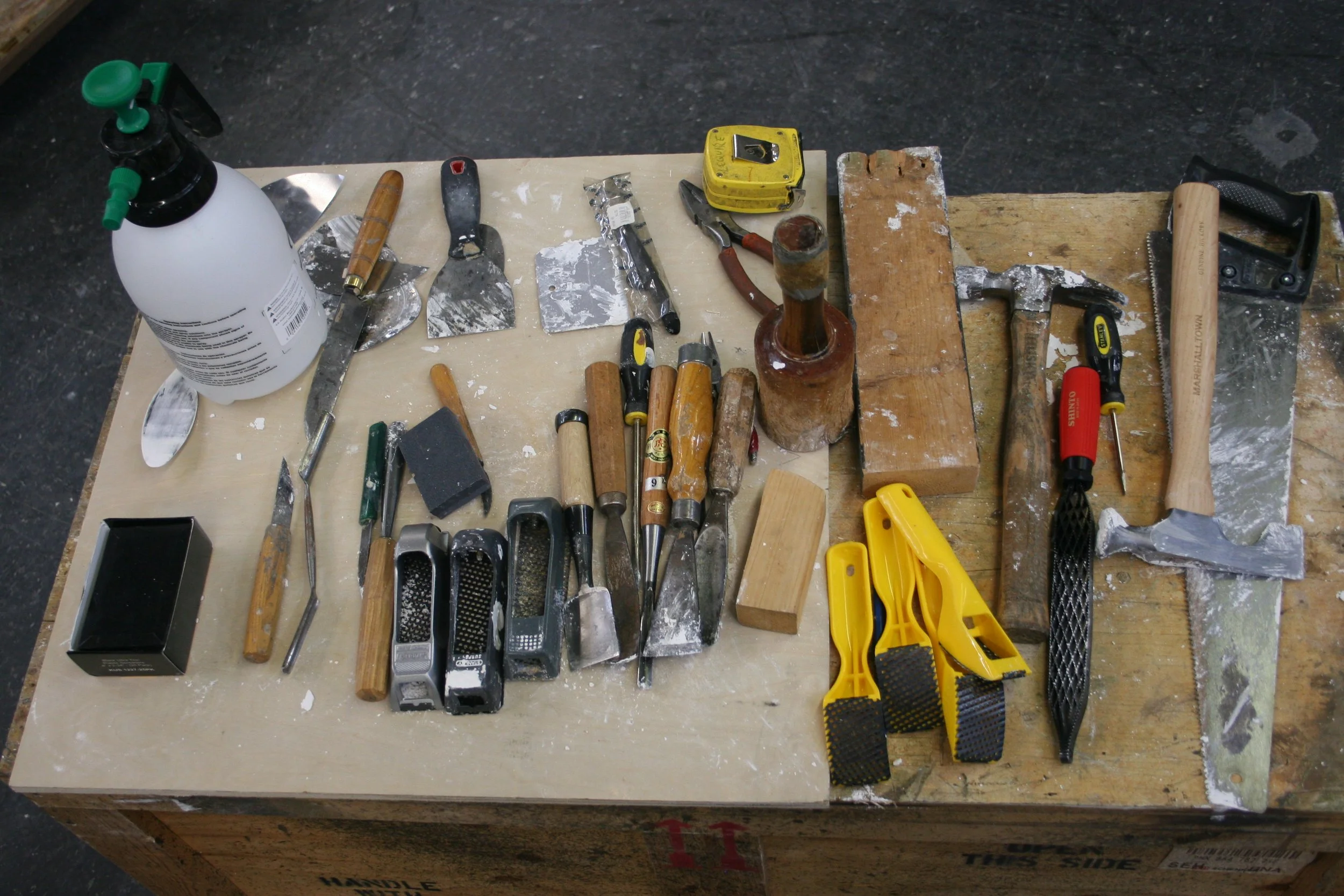 A cluttered workbench with various tools including screwdrivers, pliers, a saw, a hammer, and a bottle, all covered with dust and debris.