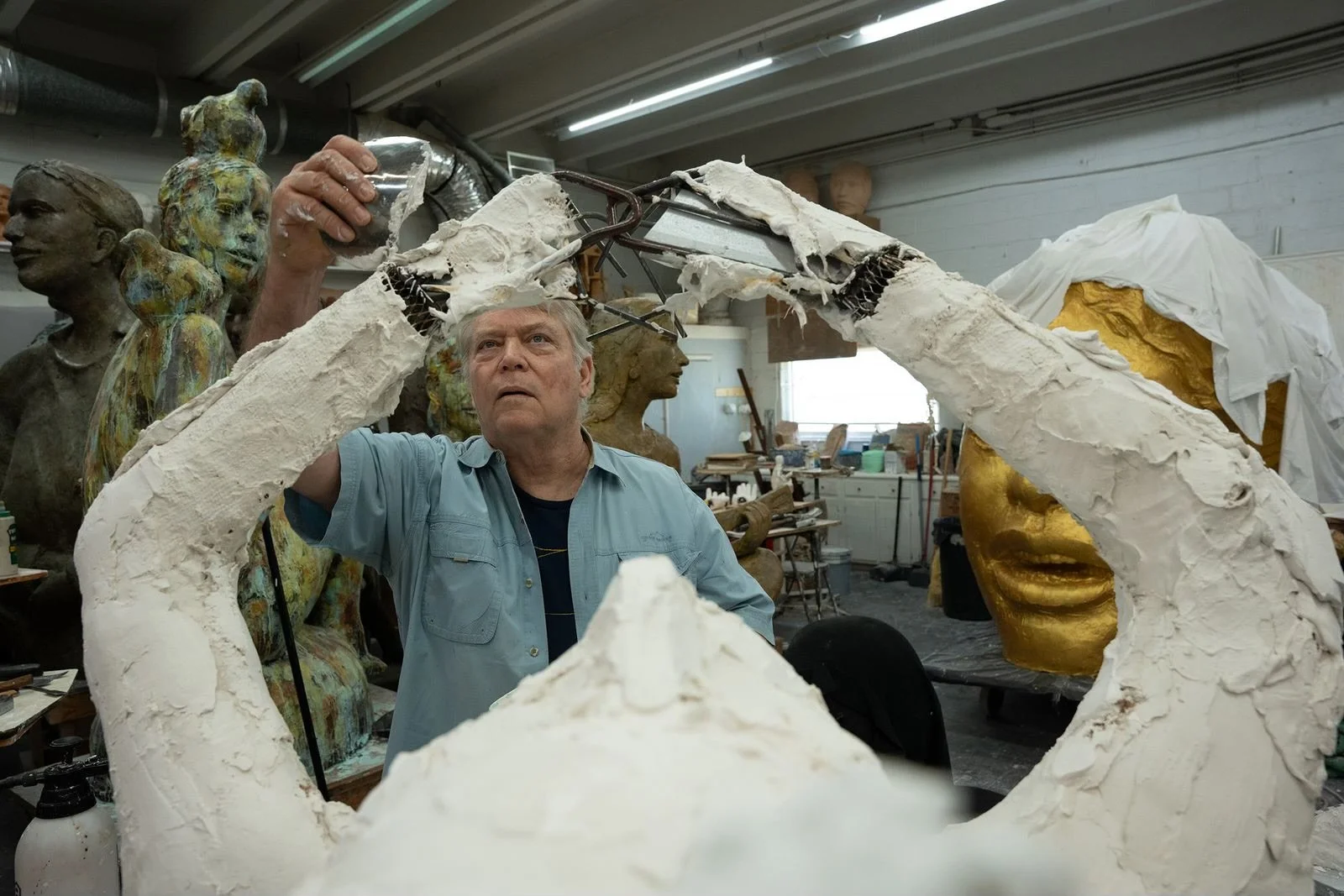 An artist working on sculpture in a studio filled with various busts and sculptures in different stages of completion.