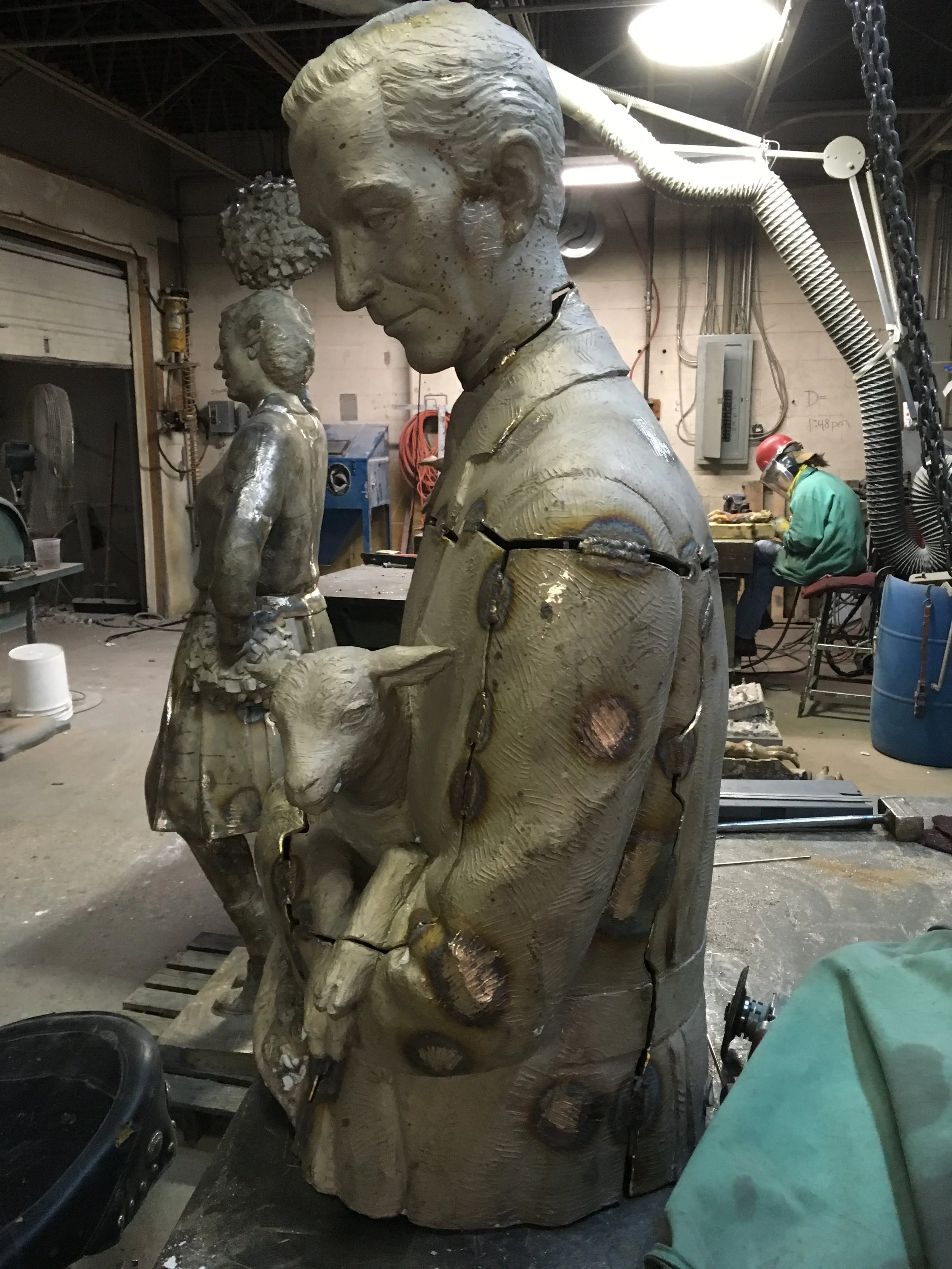 A bronze sculpture of a man and a sheep, with other sculpture projects and tools visible in a workshop environment.