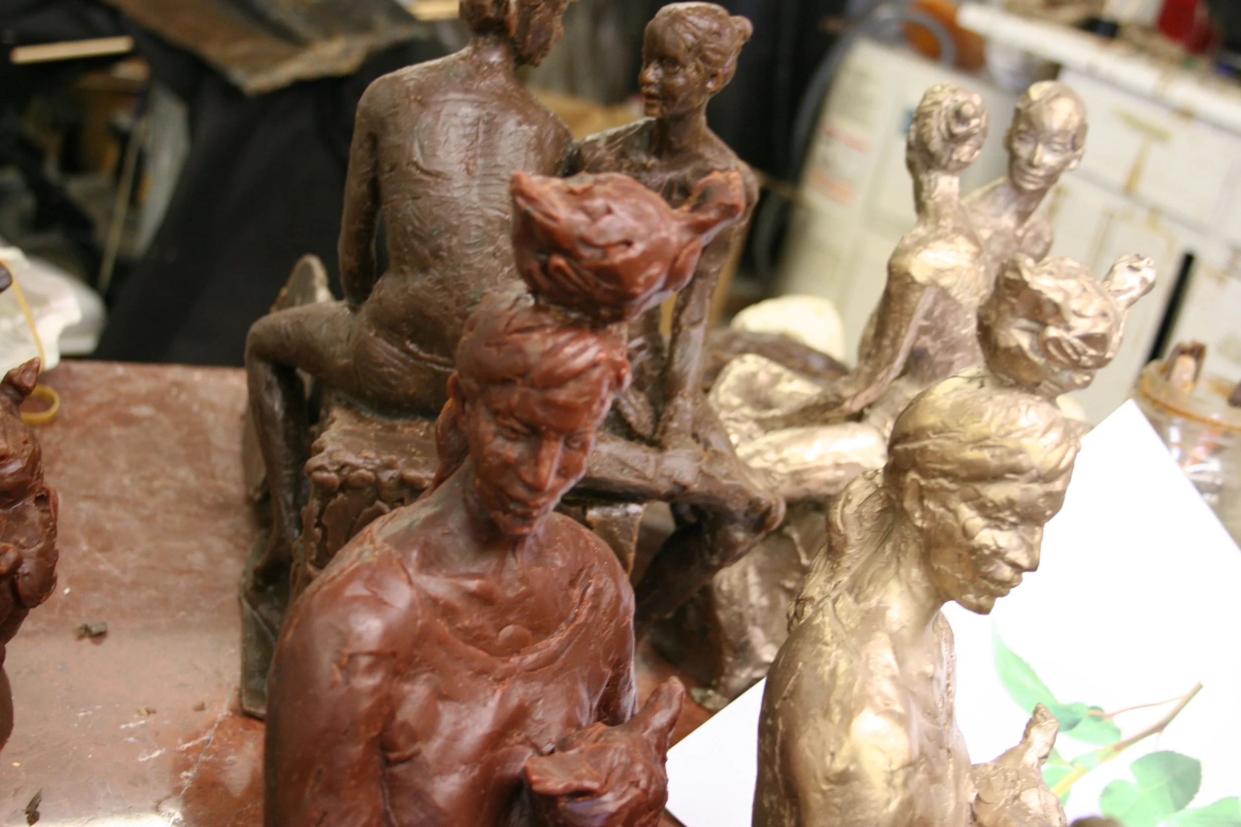 Close-up of bronze sculptures depicting a man and a woman sitting opposite each other, with two women sculpted in a different style in the background.