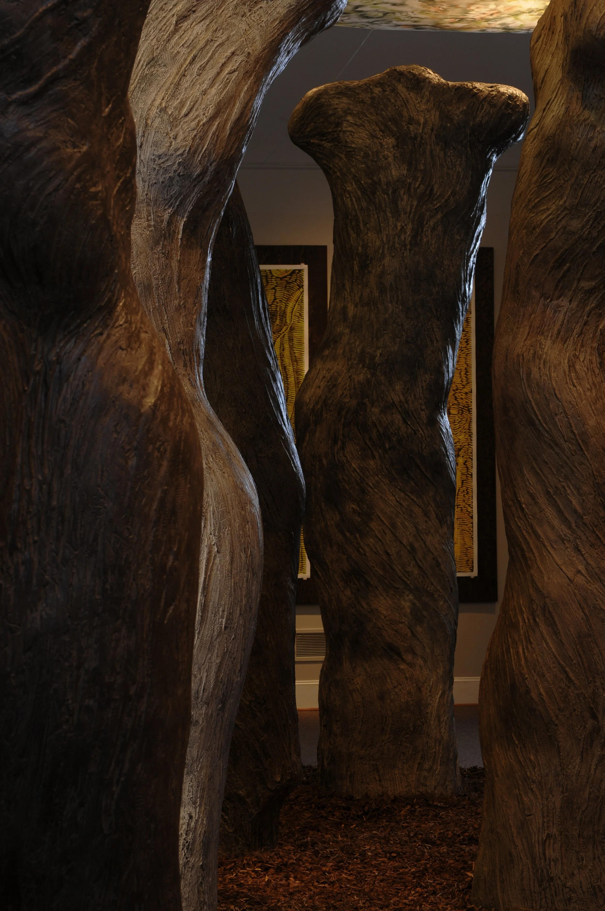 Tall wooden sculptures resembling tree trunks, displayed indoors with artwork in the background.