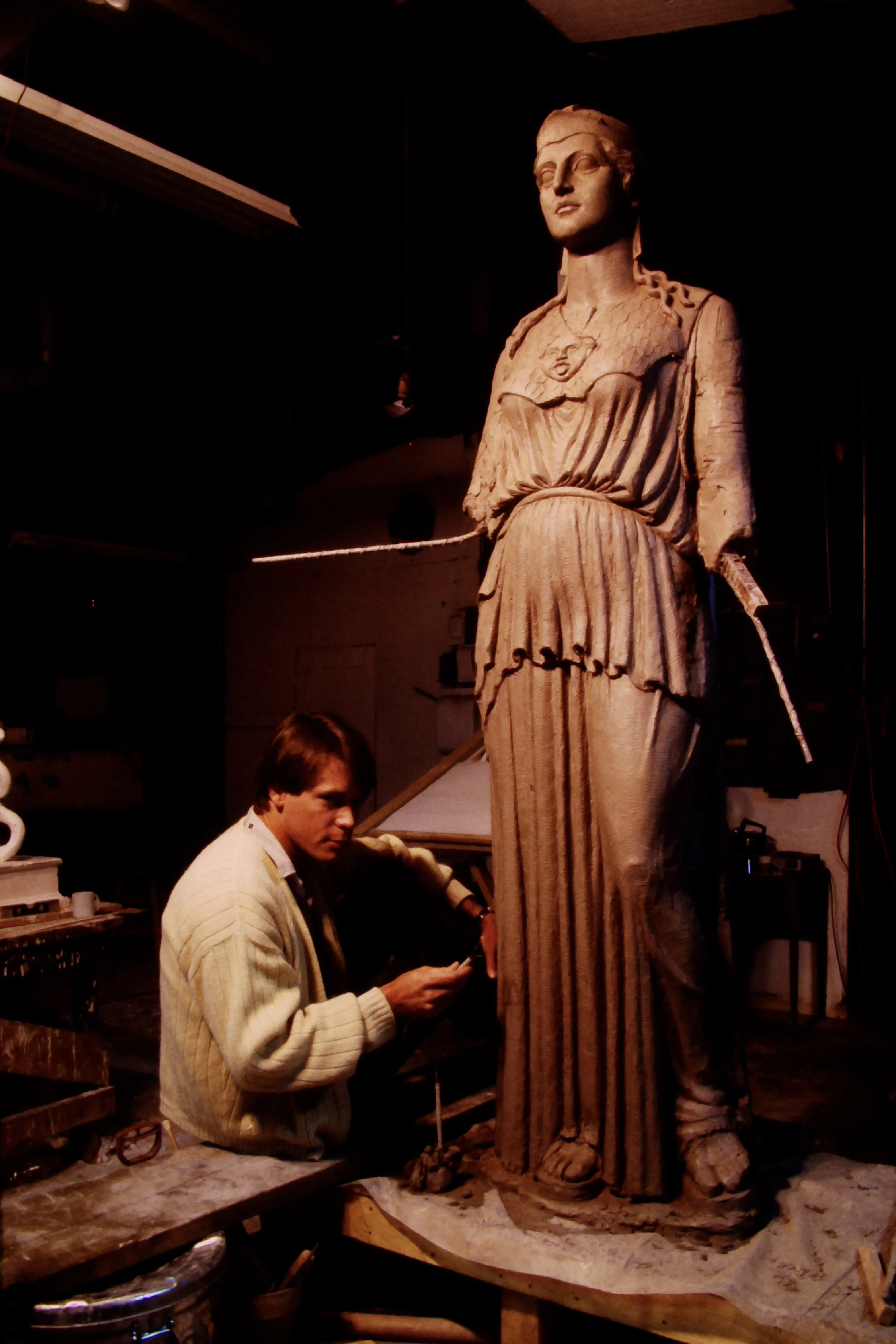 A male sculptor working on a large clay statue of a woman, with various tools and equipment around him in a workshop. Athena Parthenos in Parthenon Museum Nashville. 