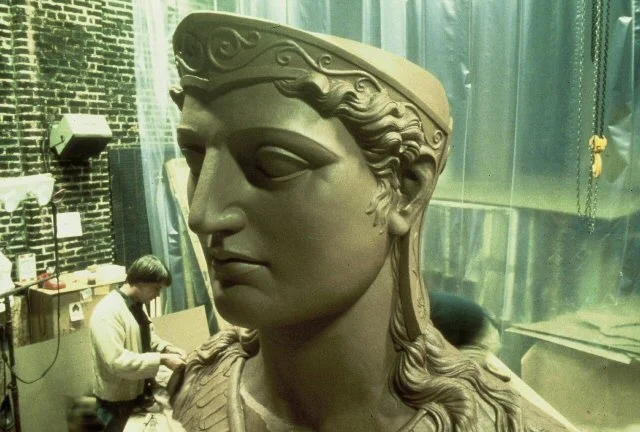 A large classical Greek style bust sculpture of a woman with intricately styled hair, in an indoor workshop or storage area, with two workers in the background. Athena Parthenos in Parthenon Museum Nashville. 