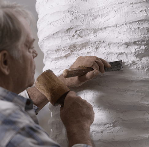Man sculpting a large white clay or plaster sculpture with a carving tool.