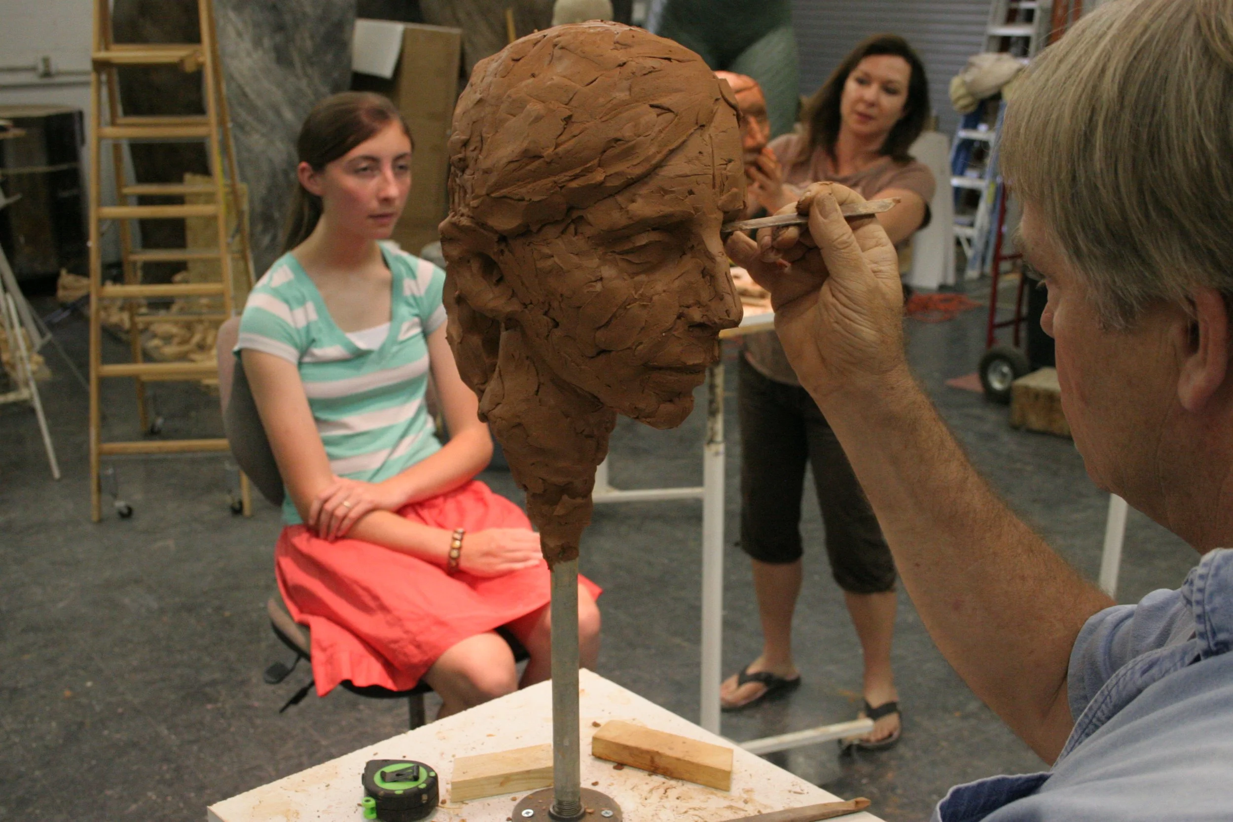 A man is sculpting a large clay bust of a person's head on a rotating stand. A woman is sitting nearby watching the process, with her arms crossed and an attentive expression. In the background, another woman is taking a photo of the sculpting proces