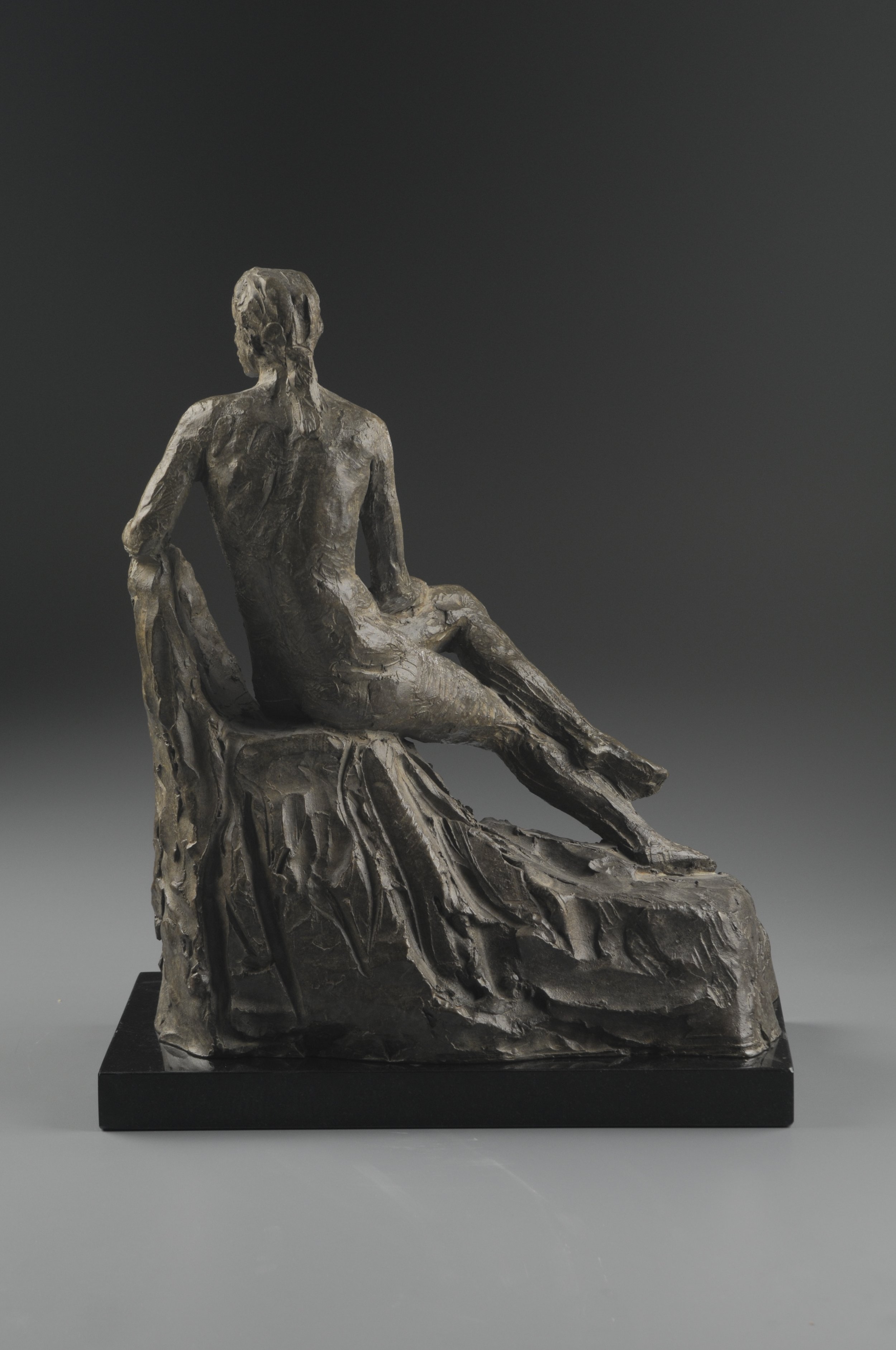 Sculpture of a seated, semi-nude man with long hair and beard on a textured base, displayed on a black platform.