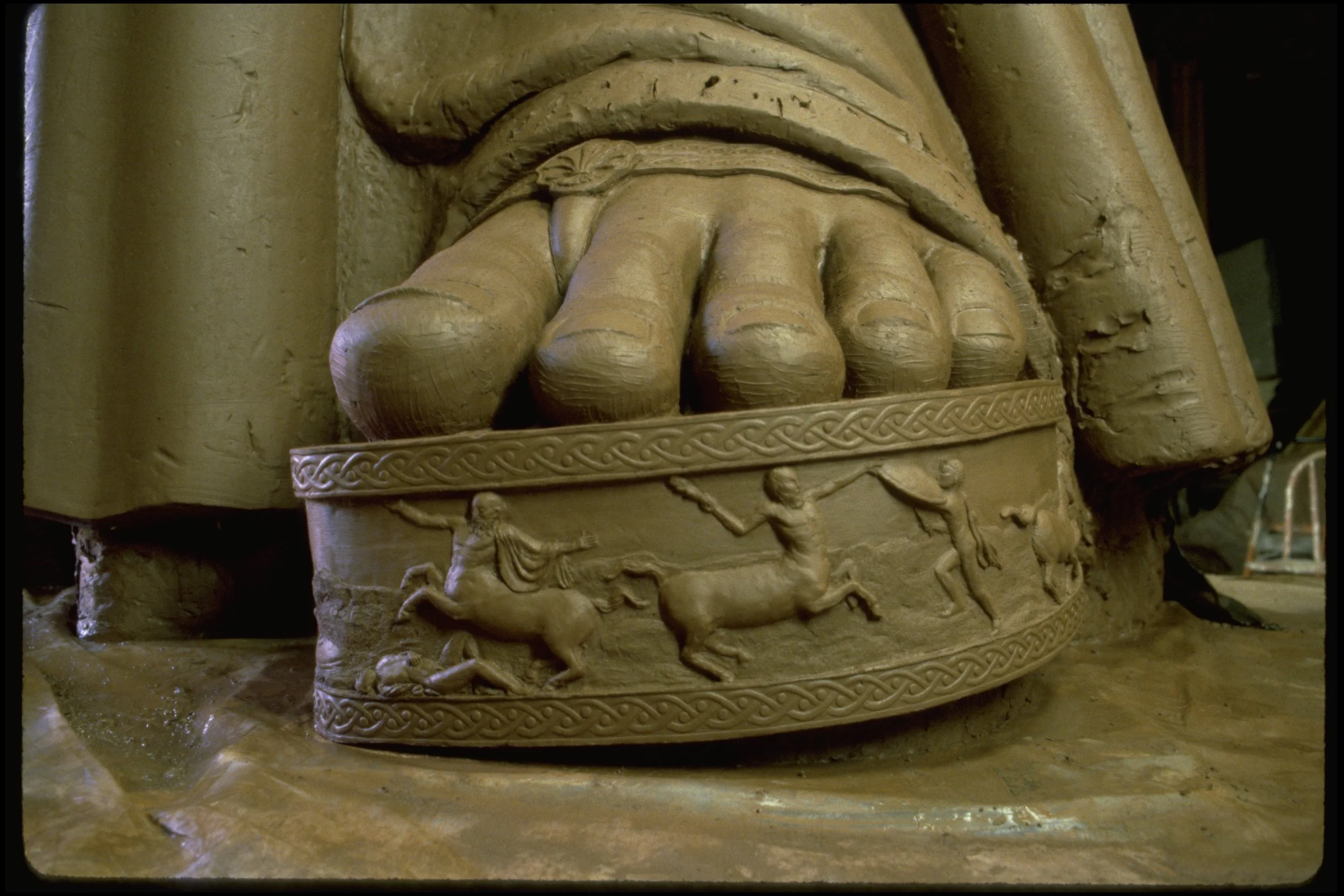Close-up of a carved clay sculpture depicting a foot with detailed toes, part of a larger decorative relief featuring intricate patterns and figures. Athena Parthenos in Parthenon Museum Nashville. 