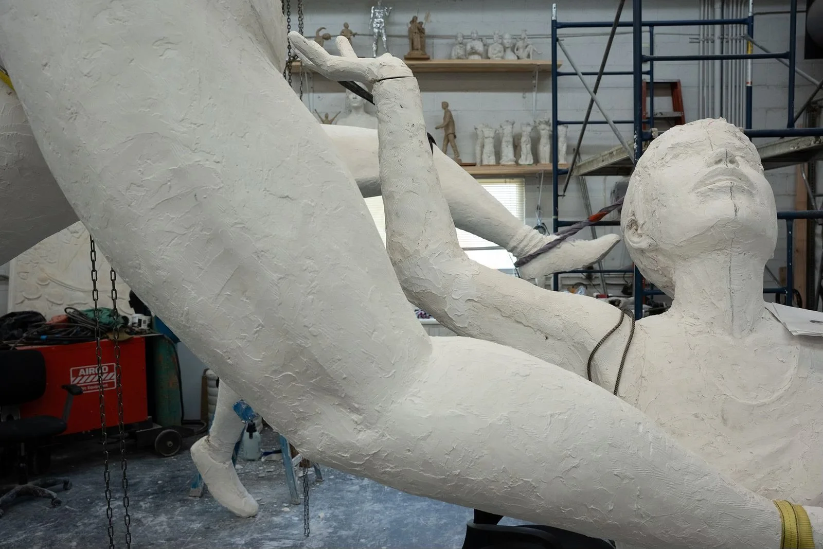A person building a large white sculpture that depicts a reclining woman with a head resting on one arm. The sculpture appears to be in the process of being formed with a textured surface. It is located in a workshop with shelves and scaffolding in t