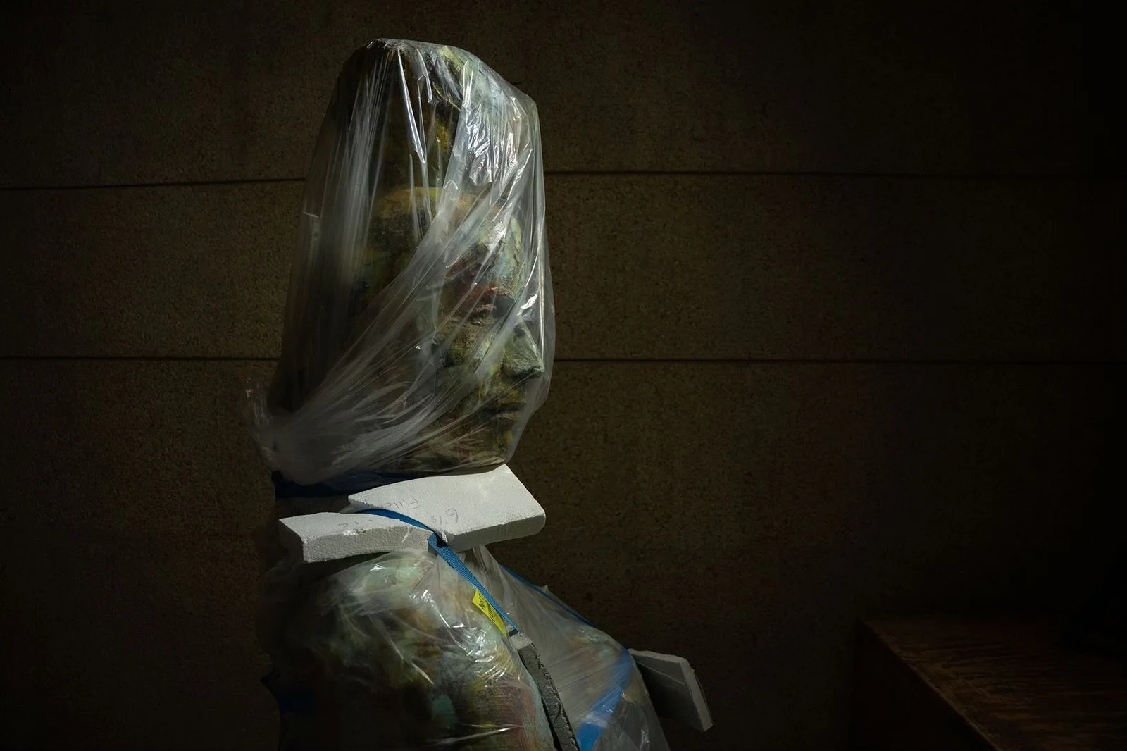 A plastic bag containing items placed on an insulated foam box, with another item underneath, set against a dark textured wall.