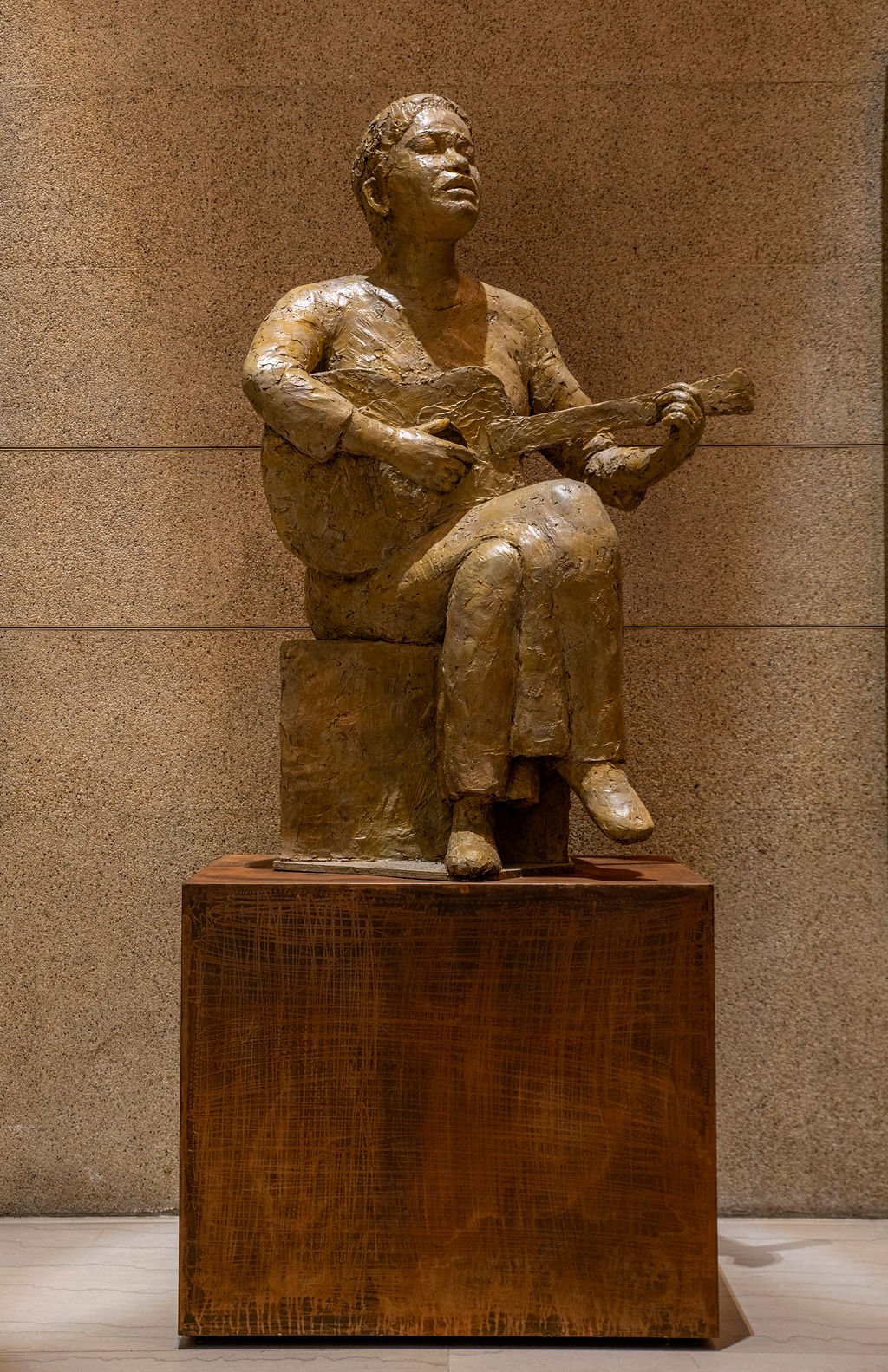 A bronze sculpture of a woman sitting on a block, playing a guitar. The sculpture is mounted on a metal pedestal against a textured wall.