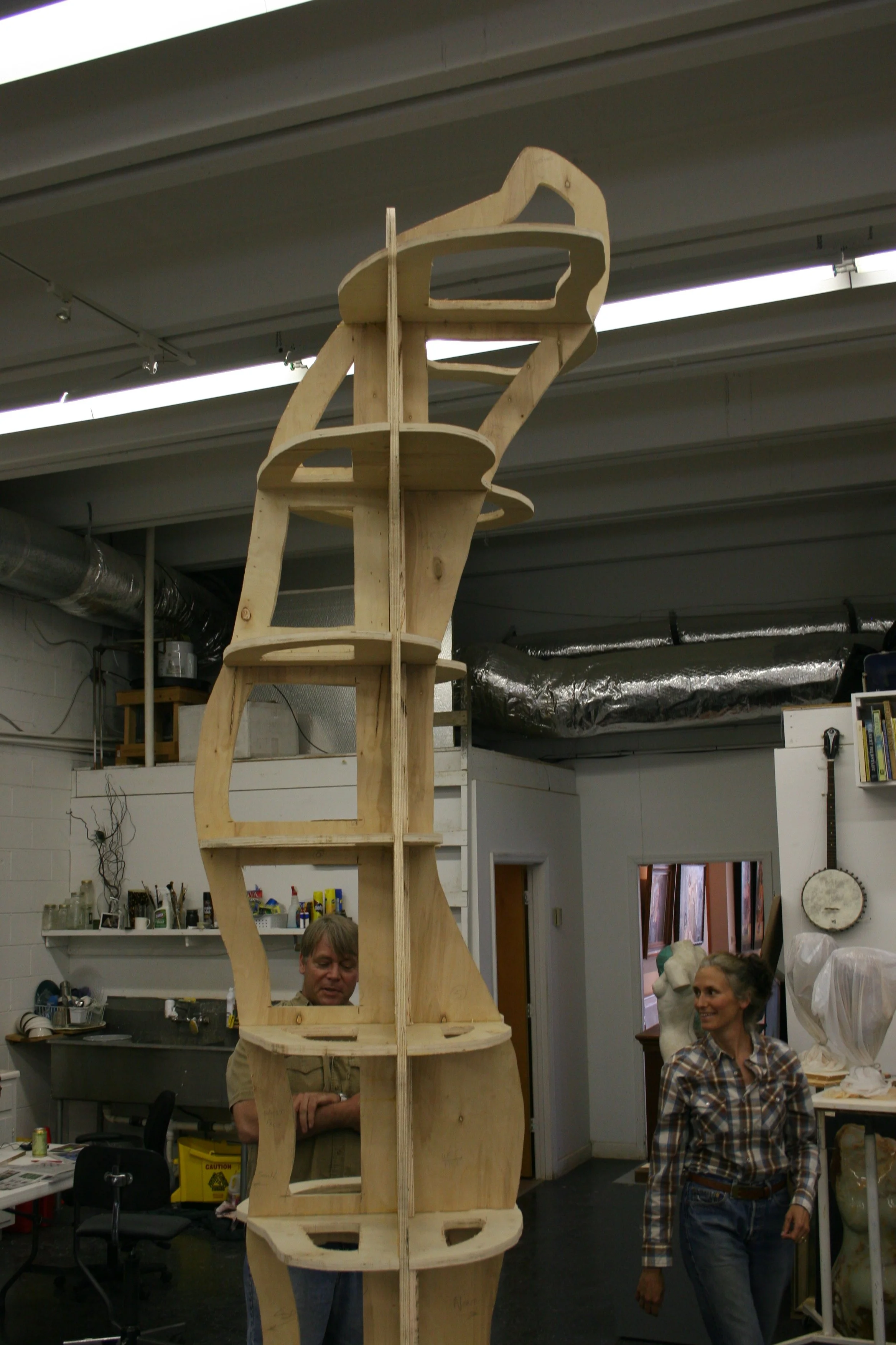 A studio with a tall wooden armature resembling a torso.. Two women are standing nearby, one with gray hair, smiling, and wearing a plaid shirt. The workshop has tools, shelves, and a skeleton model in the background.
