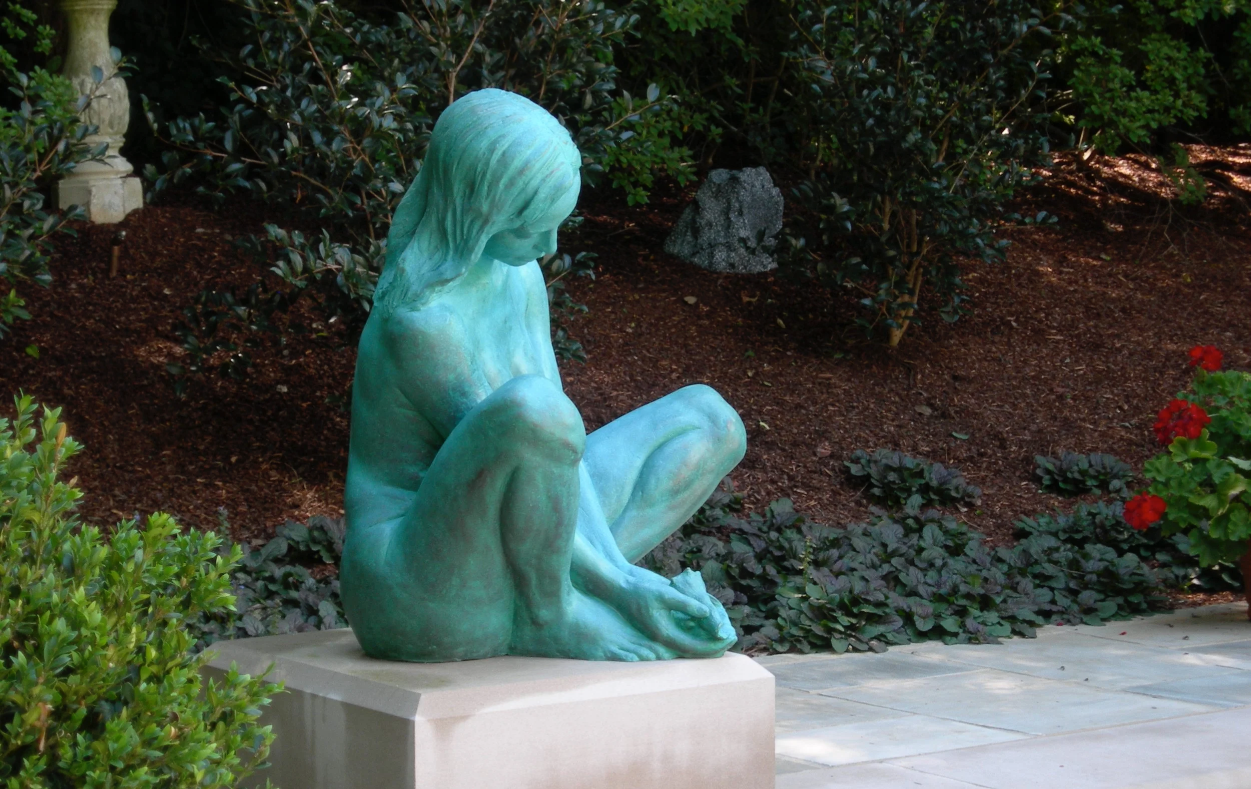 A bronze sculpture of a girl sitting with her head bowed and arms crossed over her knees, situated on a garden platform surrounded by bushes and flowers.