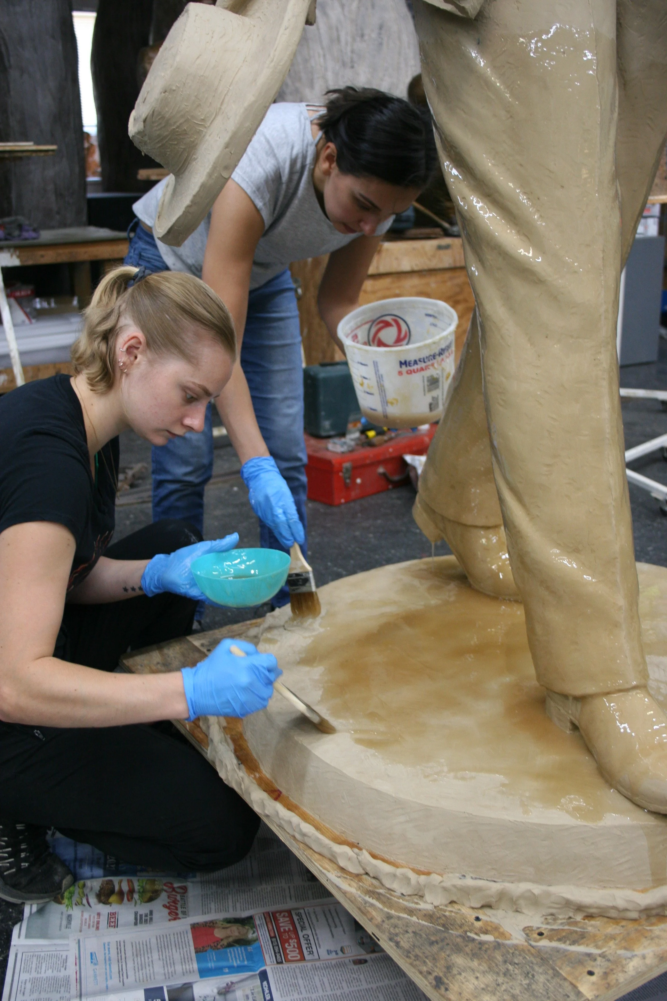 Two women working on a large sculpture of a person, applying a layer of a substance to the base of the sculpture in an art studio.