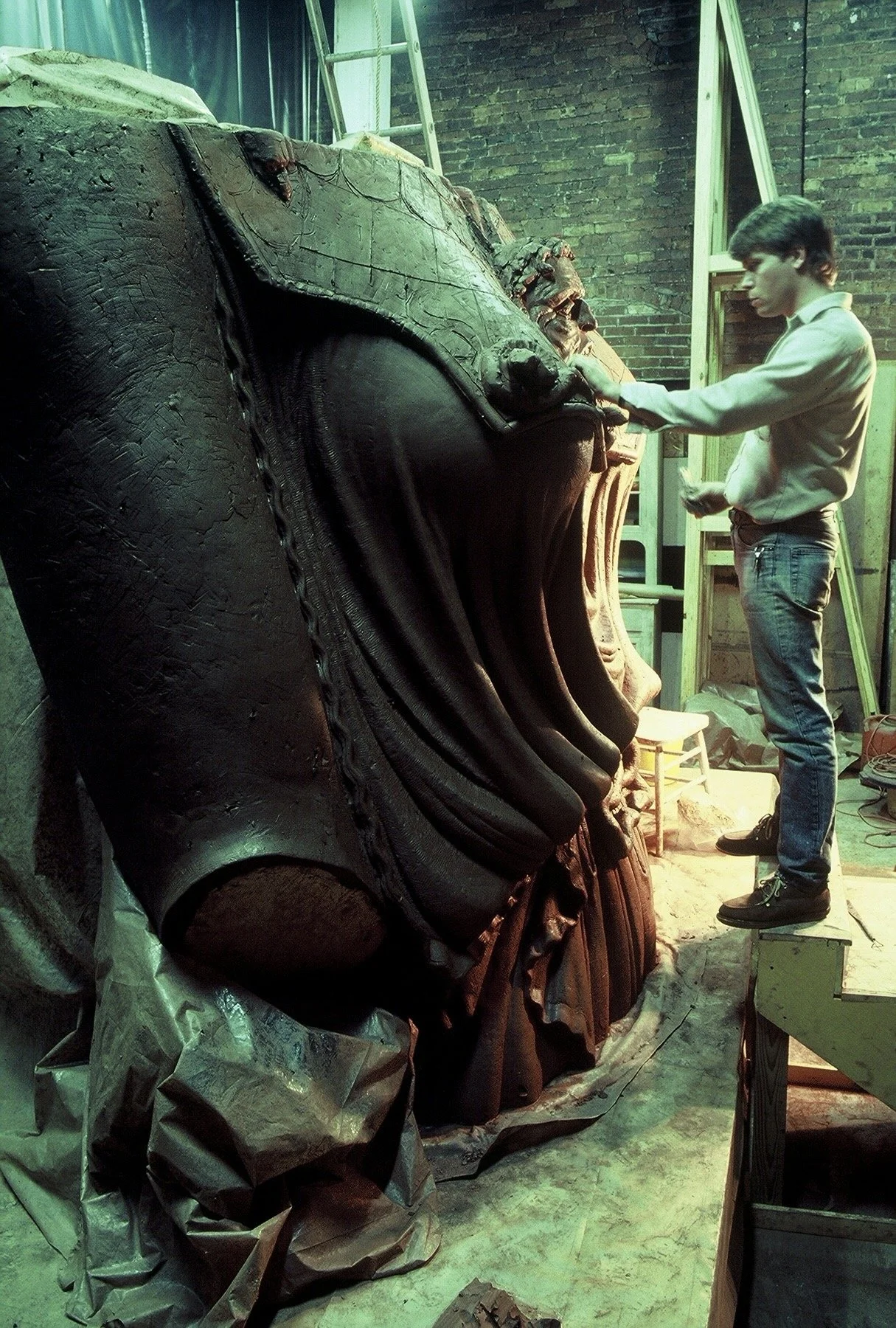 A man is standing in front of a large, detailed, sculpture in a workshop surrounded by brick walls, ladders, and workshop equipment. Athena Parthenos in Parthenon Museum Nashville. 