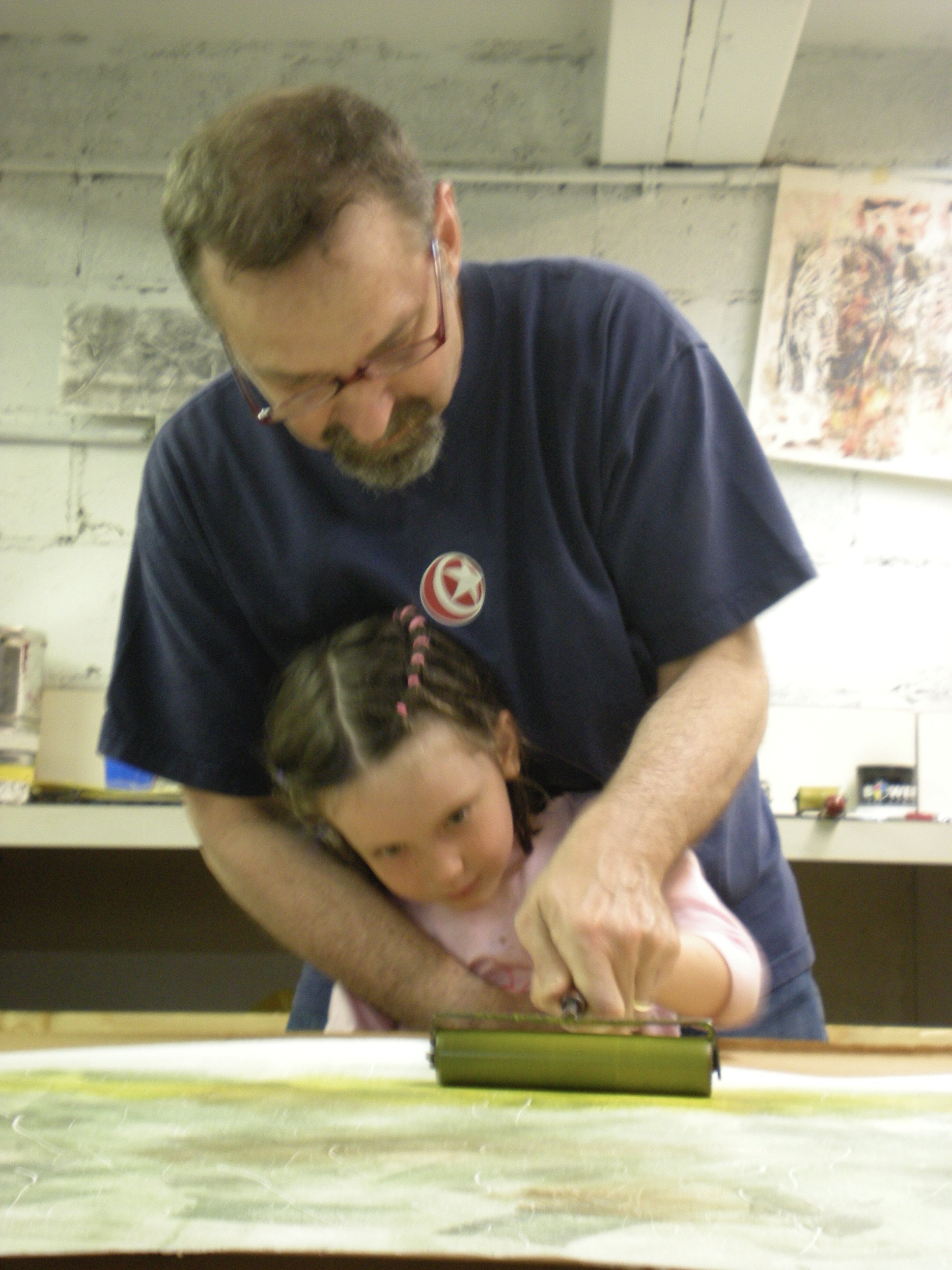An adult man with glasses and brown hair, wearing a navy blue T-shirt with a red and white logo, helping a young girl with brown hair in braids with pink beads, use a green device on a table in a workshop or art studio with art on the walls.