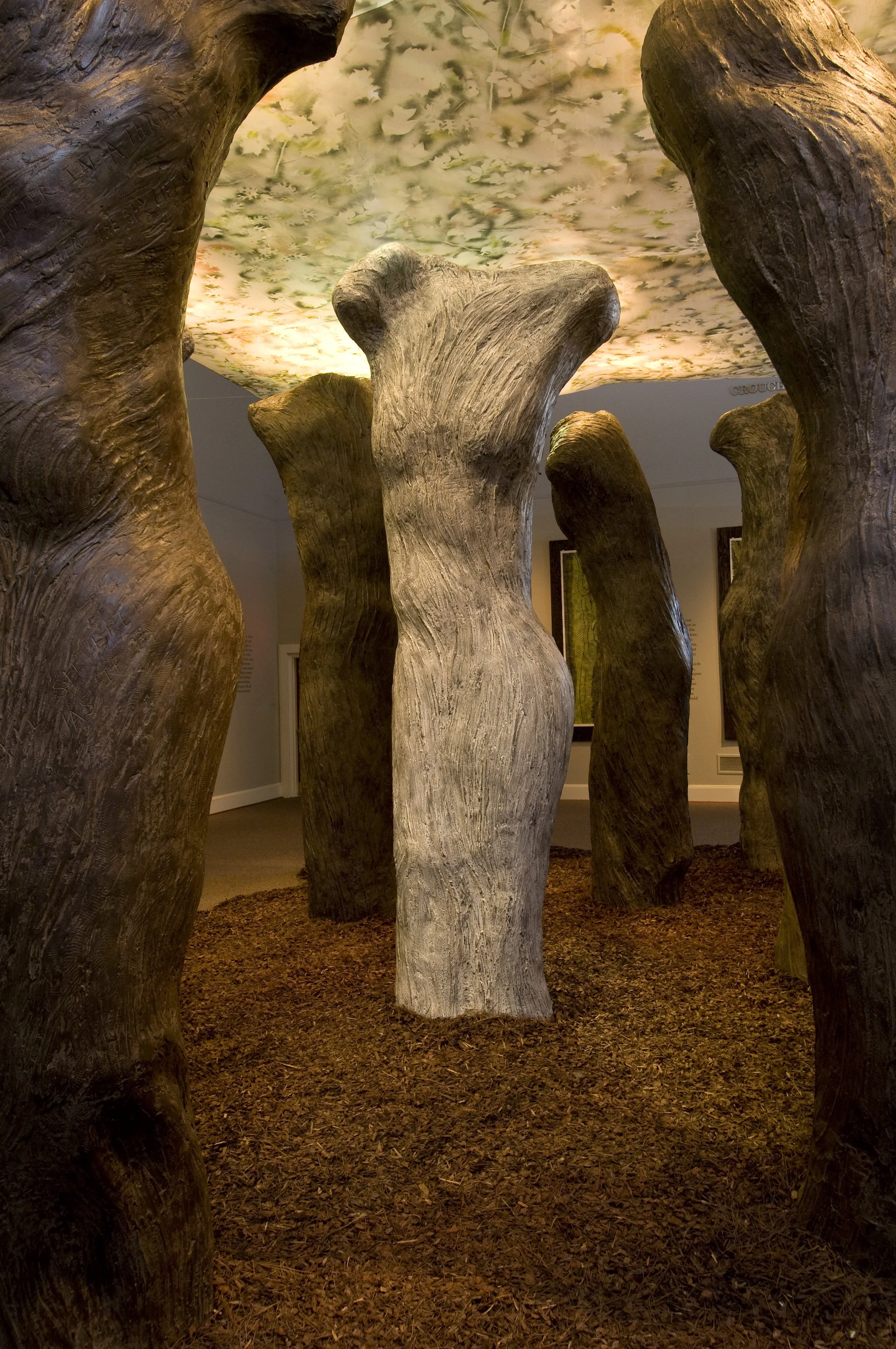 Art installation resembling a forest of large wooden tree trunks with a ceiling painted like a sky with clouds.