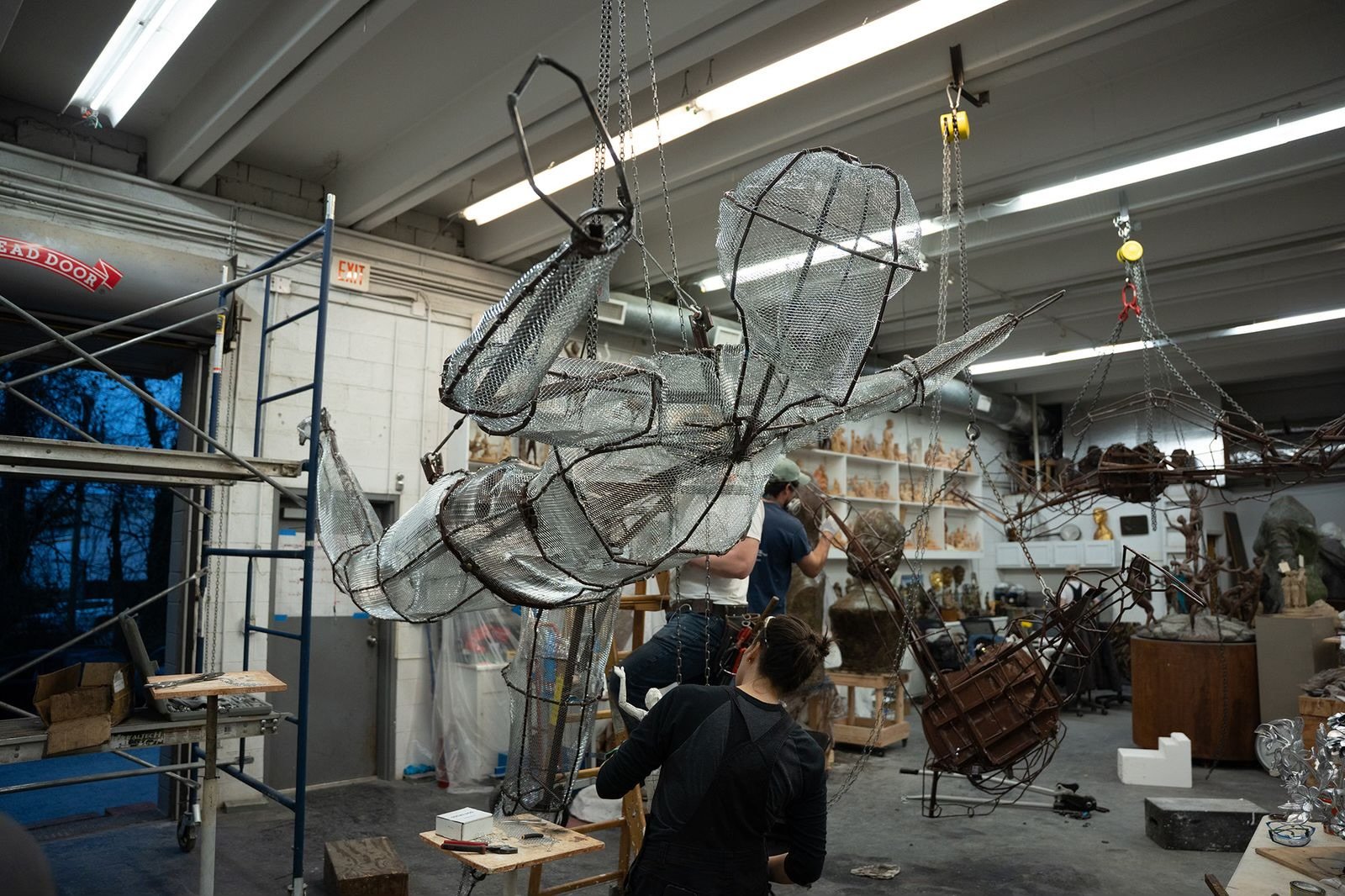 A large metallic sculpture of a person in a dynamic pose, suspended from the ceiling by chains and wires, in an art studio with shelves of sculptures and art supplies, and Amber Lelli working on various projects in the background. 